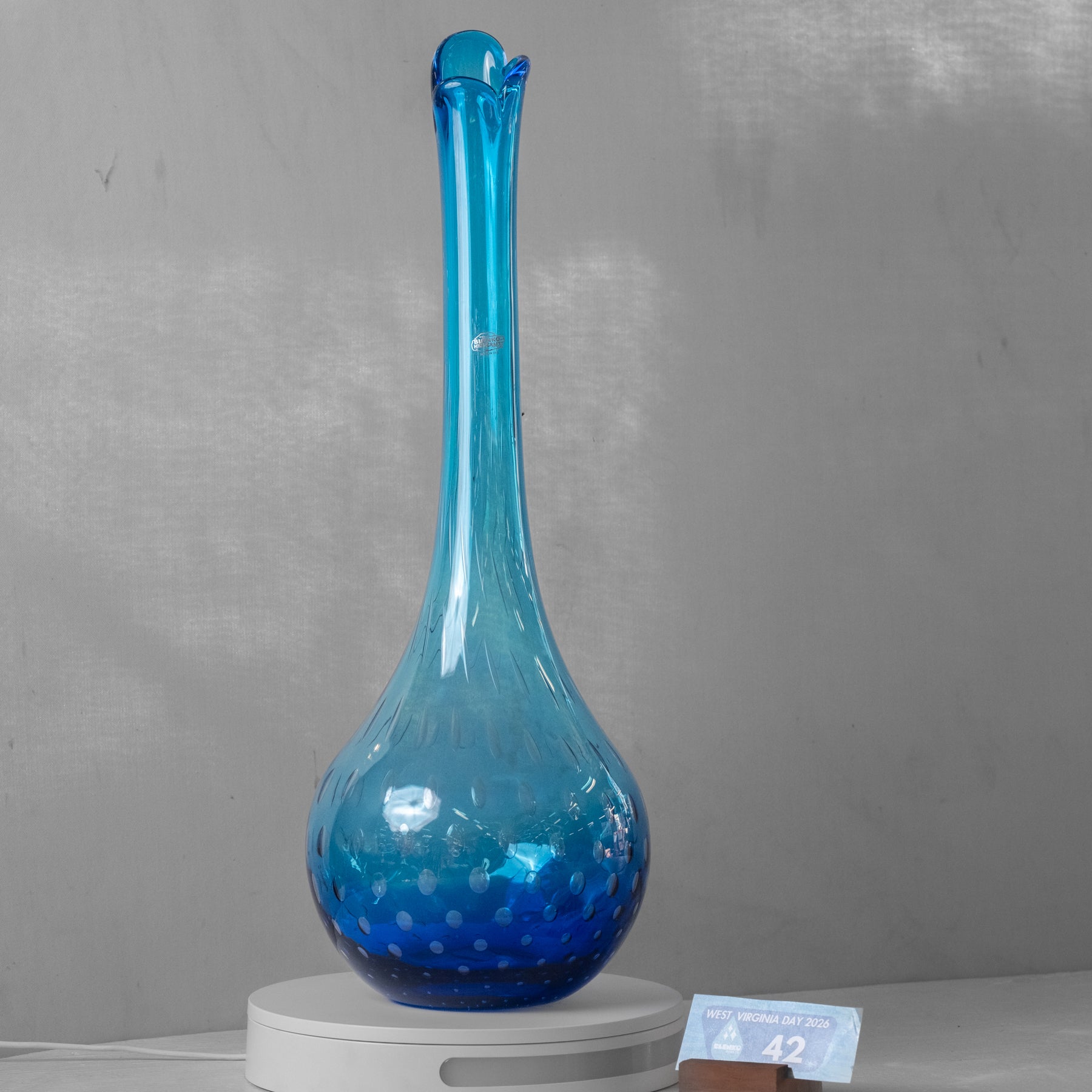 A translucent blue glass vase with a flared neck and rounded base, displayed on a white circular scale against a gray wall.
