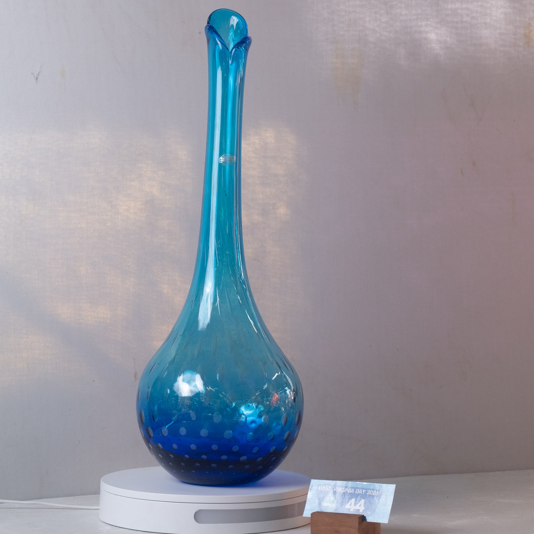 A translucent blue glass vase with a long neck and flared rim, displayed on a white circular base against a light gray wall.