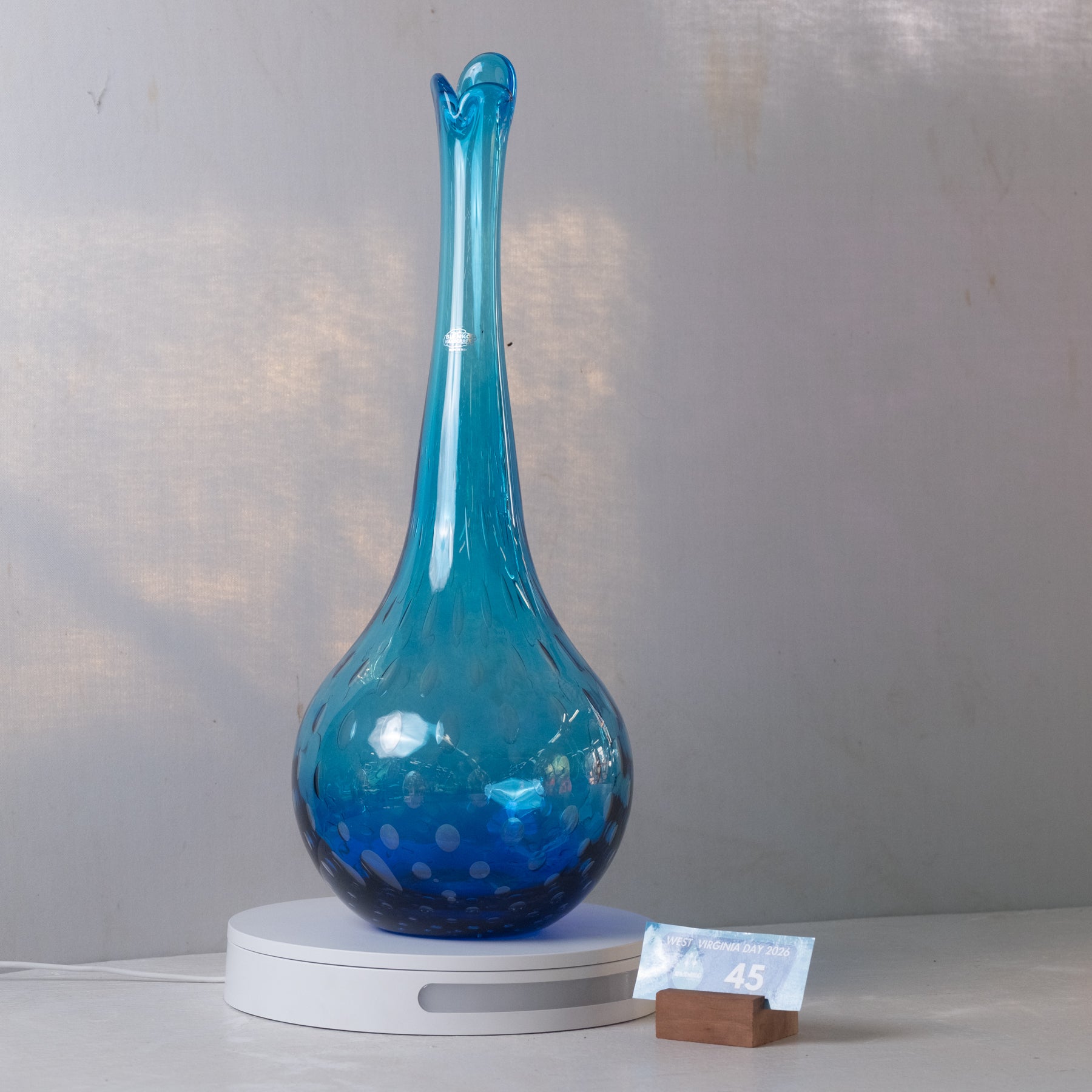 A translucent blue glass vase with a flared neck and subtle bubble pattern, displayed on a white circular light box with a small wooden block and card in front, set against a light gray wall.