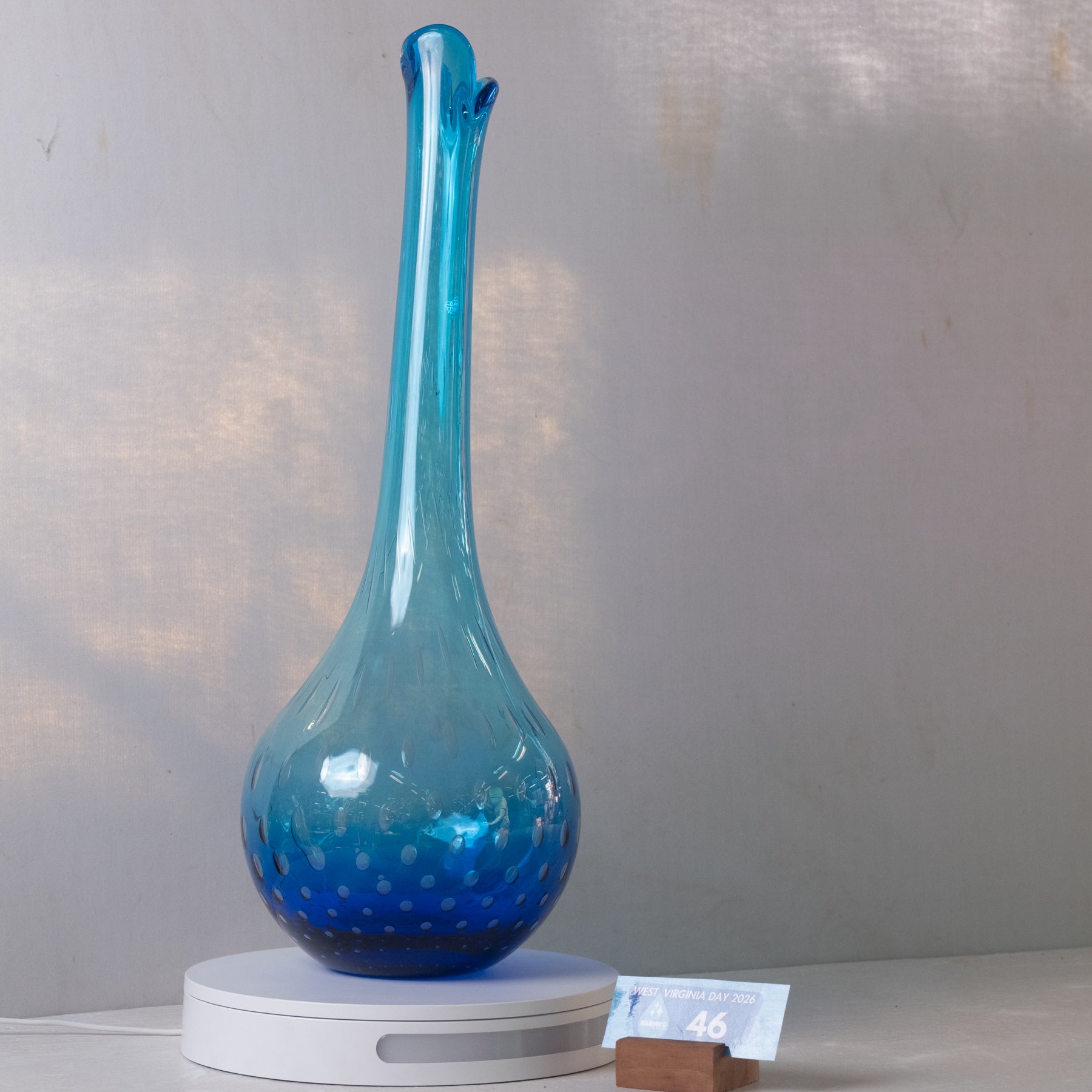 A tall, slender blue glass vase with a bulbous base and small dotted pattern near the bottom, displayed on a white circular light box against a light gray wall.