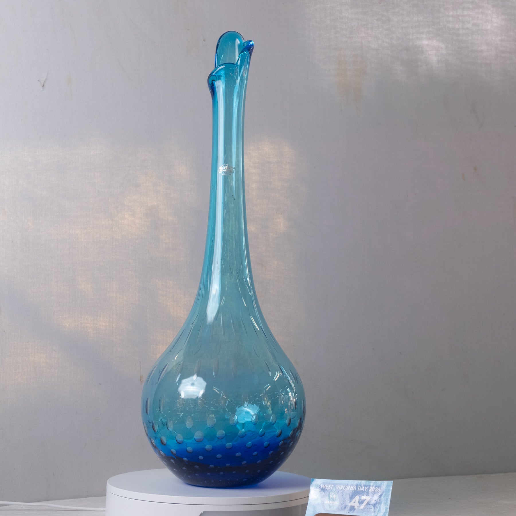 A translucent blue glass vase with a bulbous base, narrow neck, and flared rim, displayed on a white pedestal against a light gray wall.
