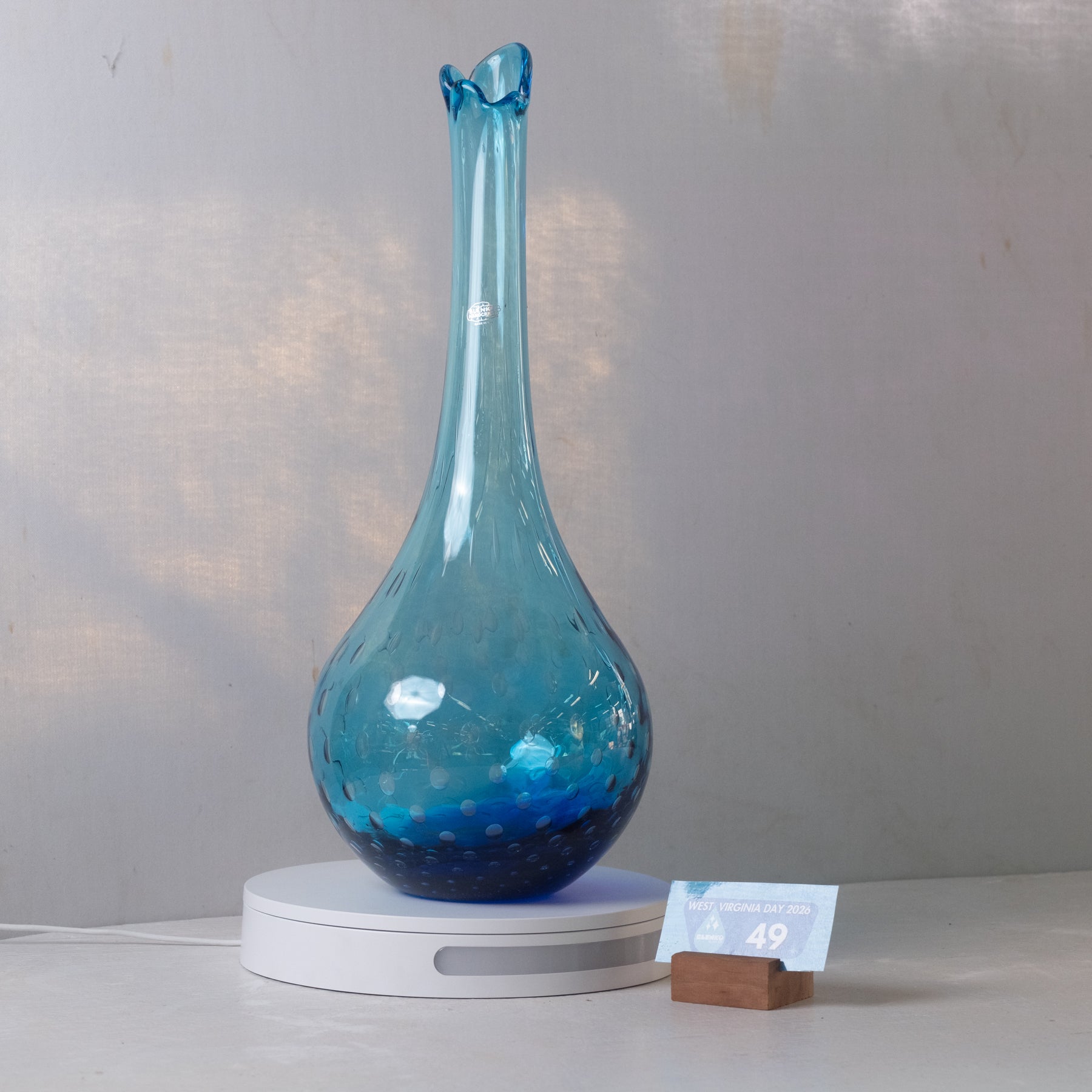 A blue glass vase with a flared neck and speckled bubble pattern, displayed on a white circular charging base against a light gray wall.