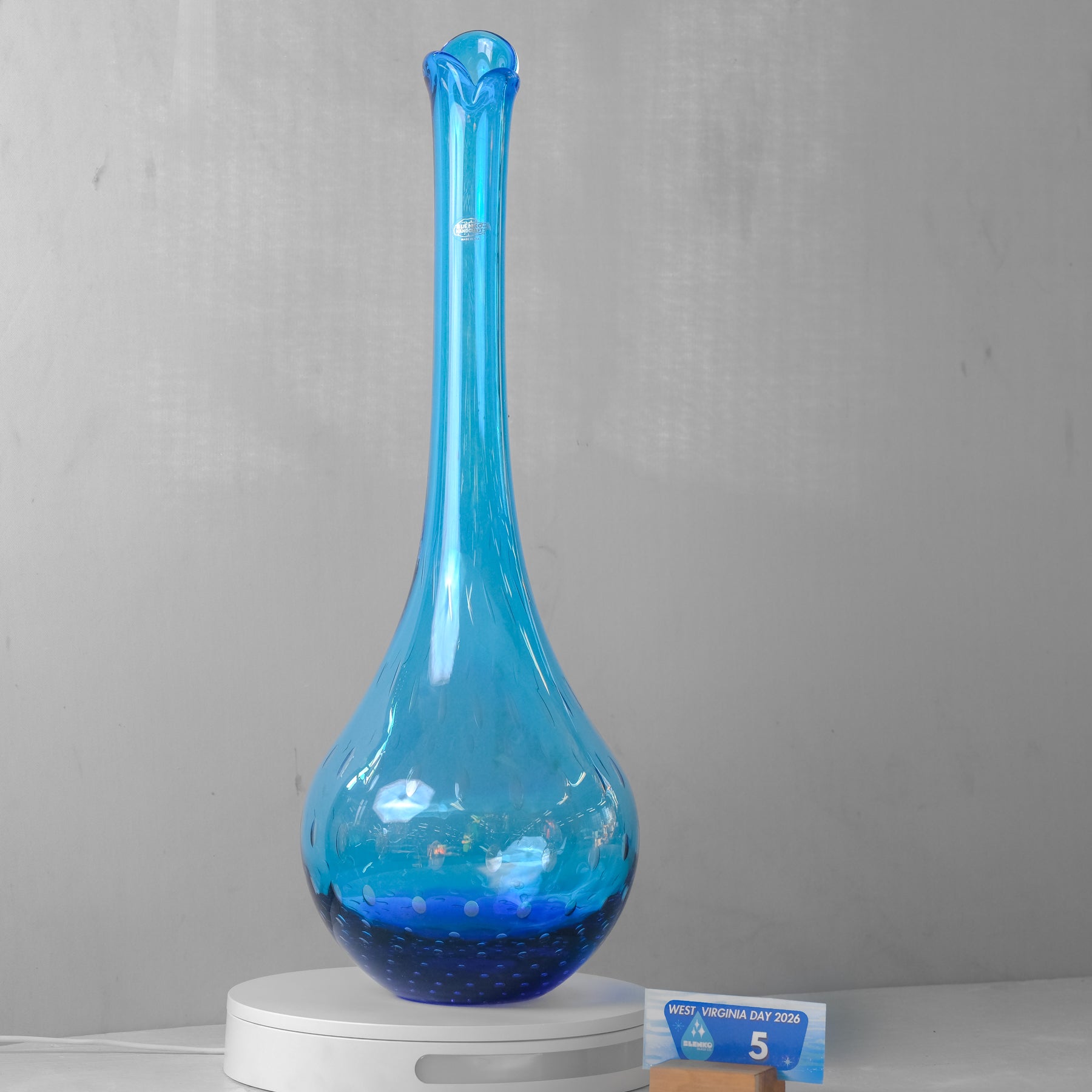 A tall, slender blue glass vase with a bulbous rounded base and a flared, ruffled neck, displayed on a white circular platform against a gray wall.