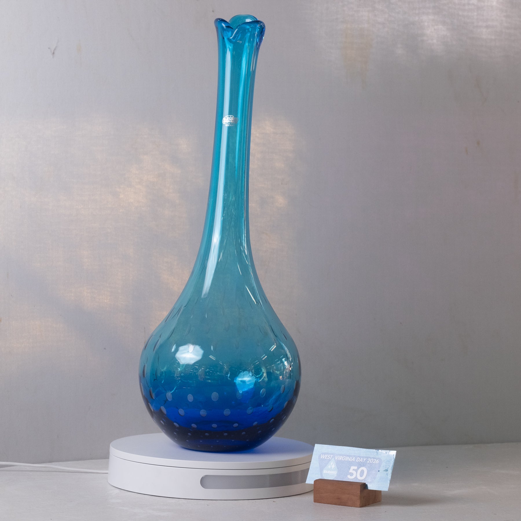 A blue glass vase with a flared neck and dotted pattern, displayed on a white circular light box with a small price tag in front, set against a neutral gray background.