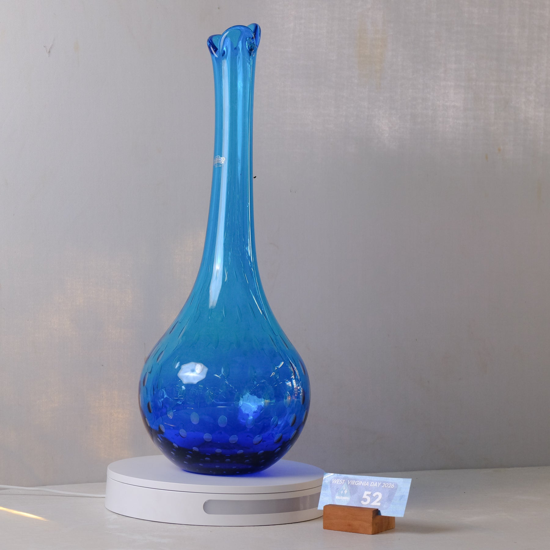 A cobalt blue glass vase with a flared, ruffled neck and a bulbous base, displayed on a white circular lighted base. A small paper price tag on a wooden block is visible in the foreground against a neutral gray textured background.