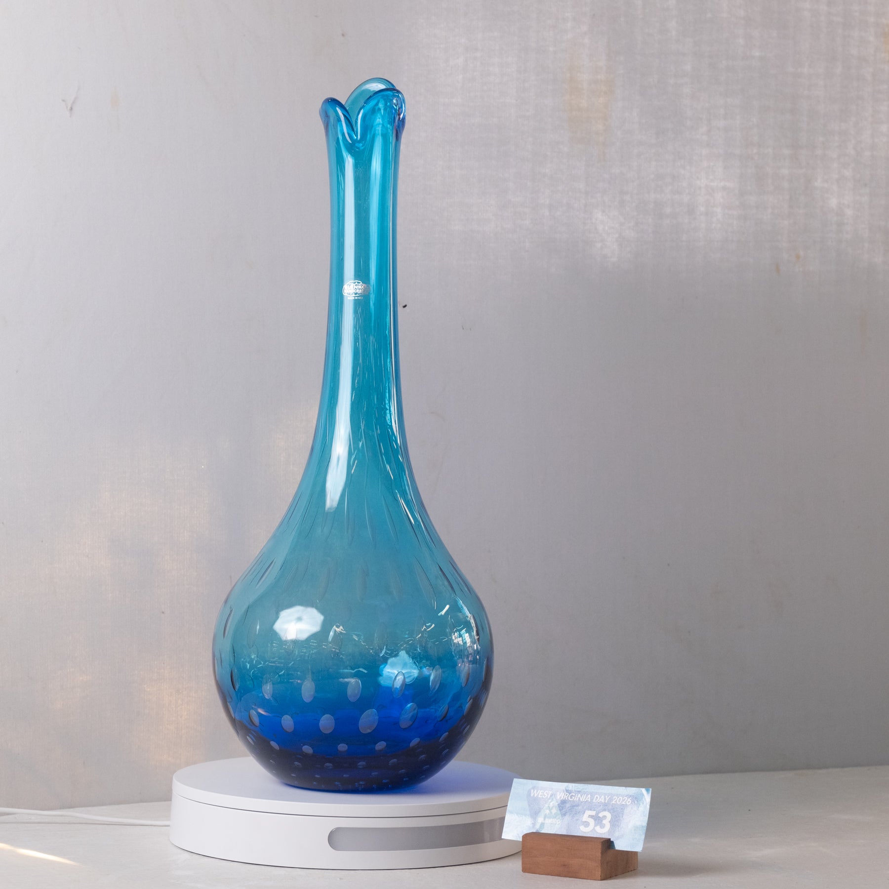 A translucent blue glass vase with a flared neck and spotted pattern near the base, displayed on a white circular lighted stand against a light gray wall.
