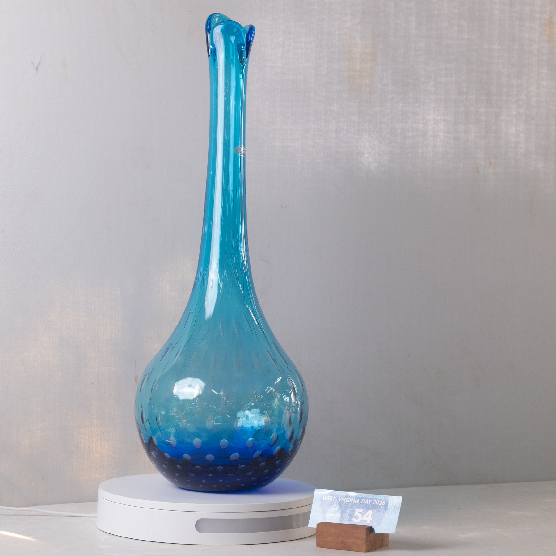 A blue glass decanter with a bulbous base and a long, flared neck, displayed on a white circular stand against a light gray textured background.