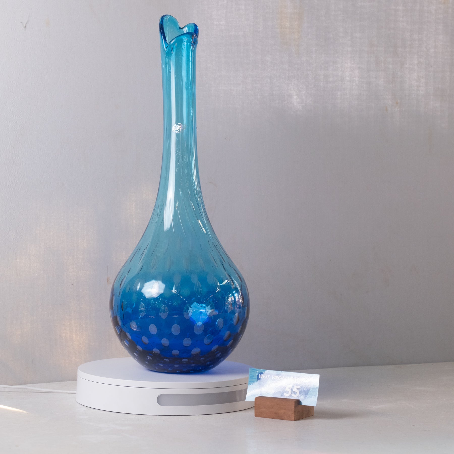 A blue glass vase with a flared neck and bubble pattern, displayed on a white circular charging base against a light gray wall.