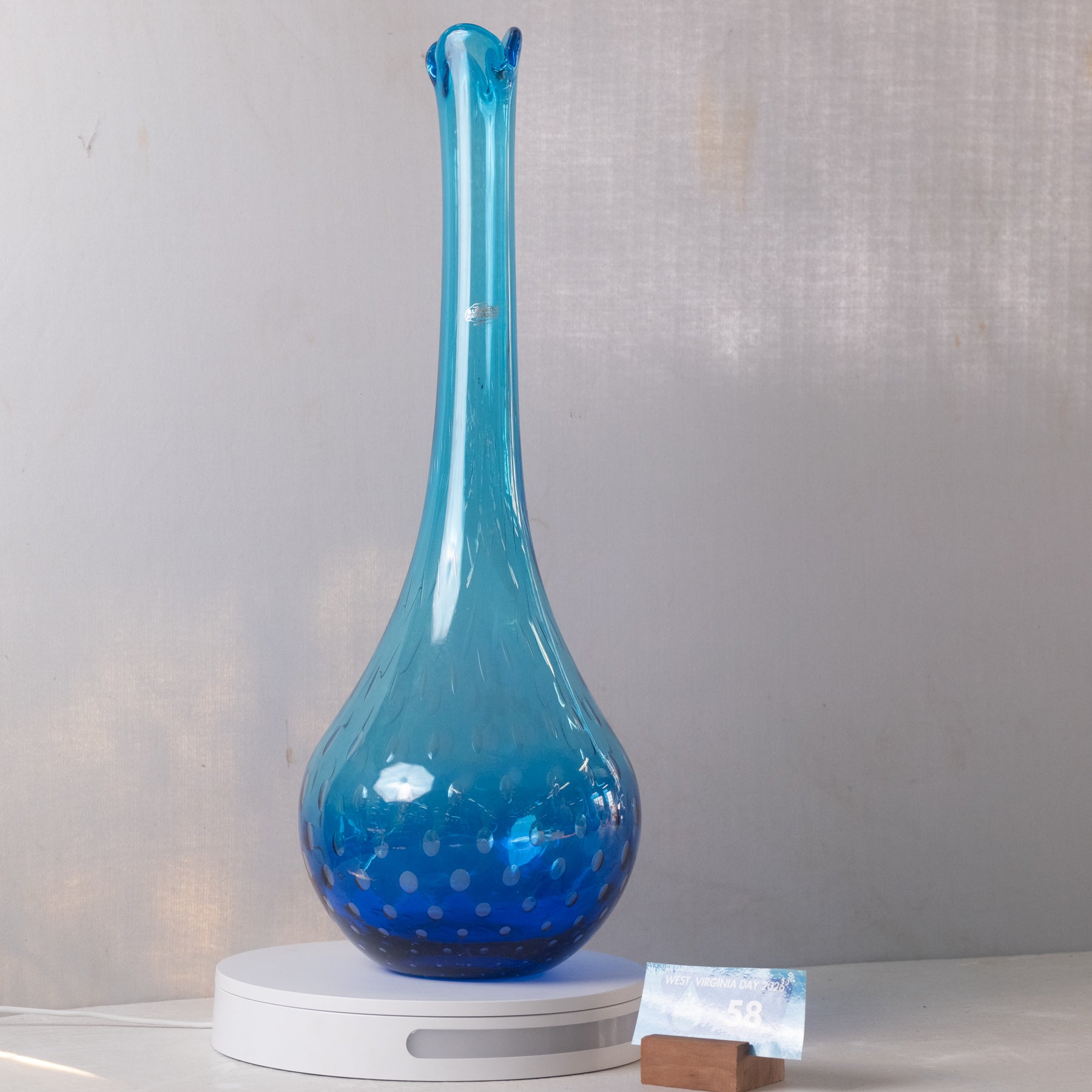 A translucent blue glass vase with a flared neck and dotted pattern near the base, displayed on a white circular stand against a light gray wall.