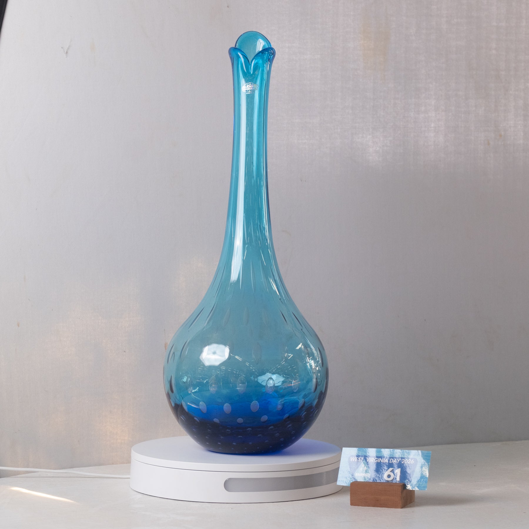 A translucent blue glass vase with a flared rim and bulbous base, displayed on a white circular light box. A small wrapped item sits beside the base against a light gray backdrop.
