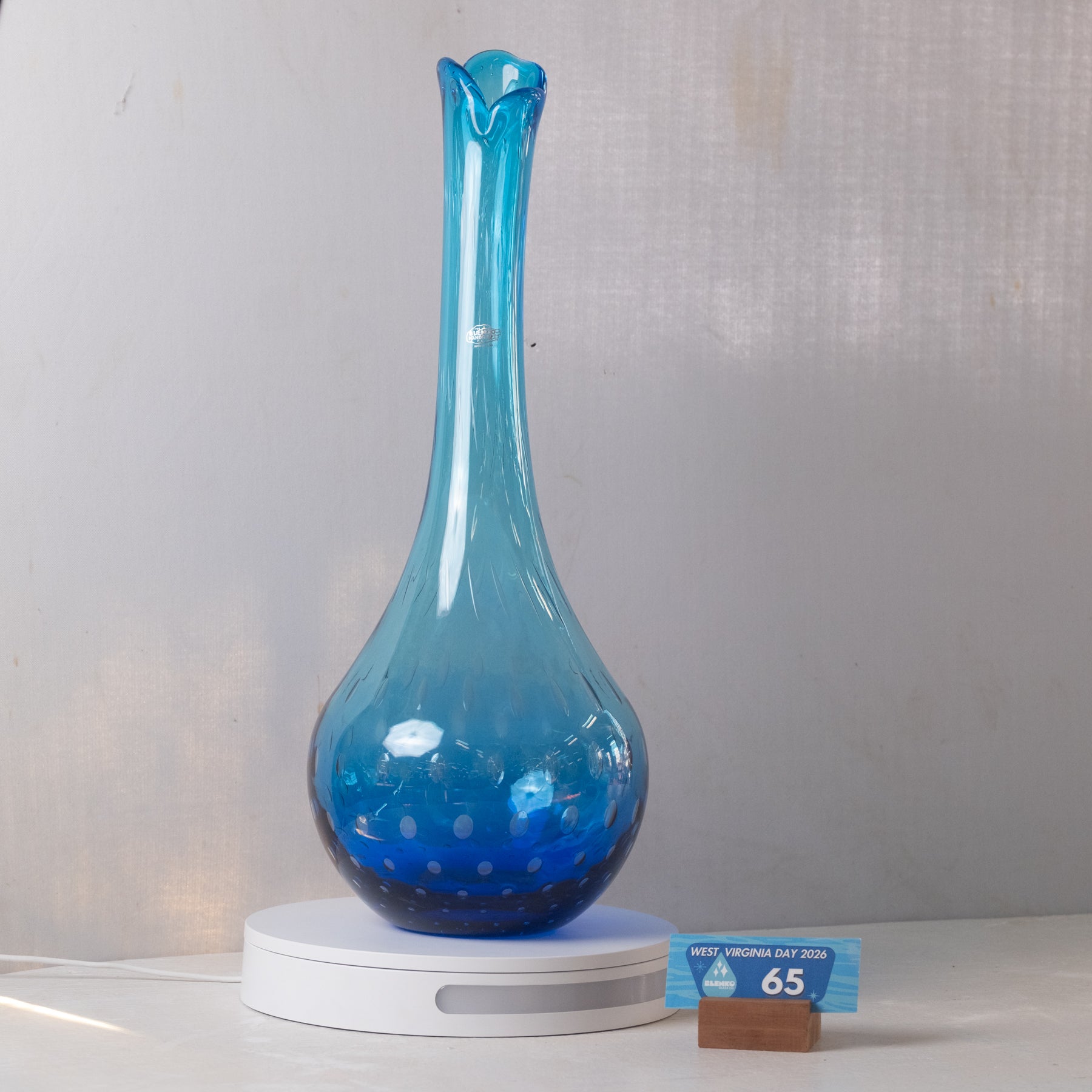 A blue glass vase with a flared neck and dotted pattern near the base, displayed on a white circular lighted base against a light gray fabric backdrop.