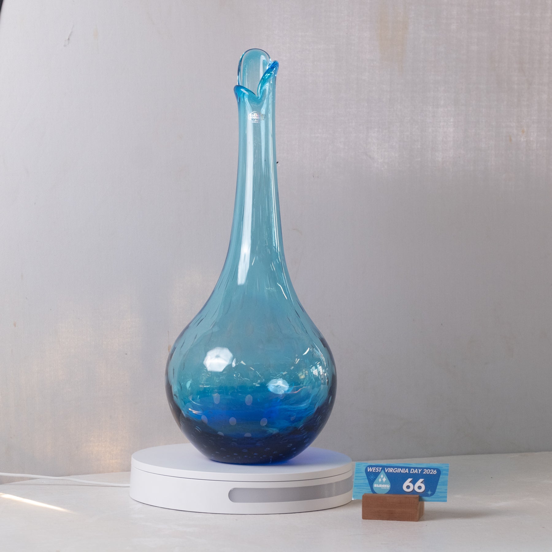 A translucent blue glass vase with a bulbous base and narrow flared neck, displayed on a white circular stand against a light gray backdrop. A small blue price card on a wooden block sits to the right.