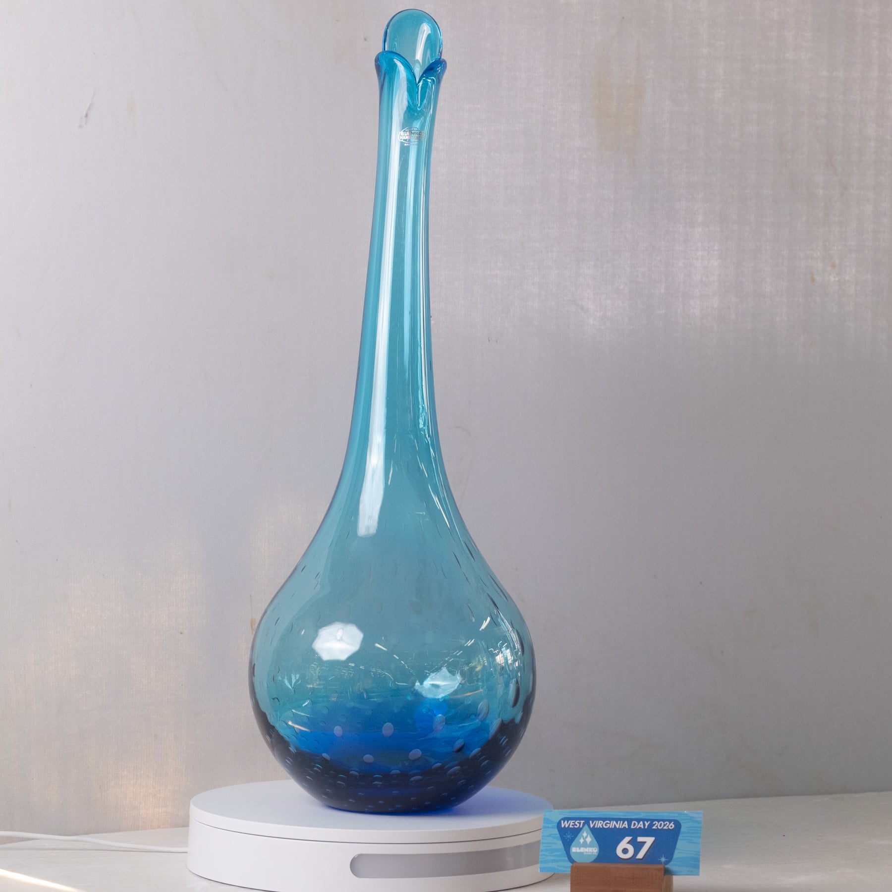 A tall, slender blue glass decanter or vase with a bulbous base, displayed on a white circular light box against a light gray fabric backdrop. A small blue and brown price tag card is visible in the lower right corner.