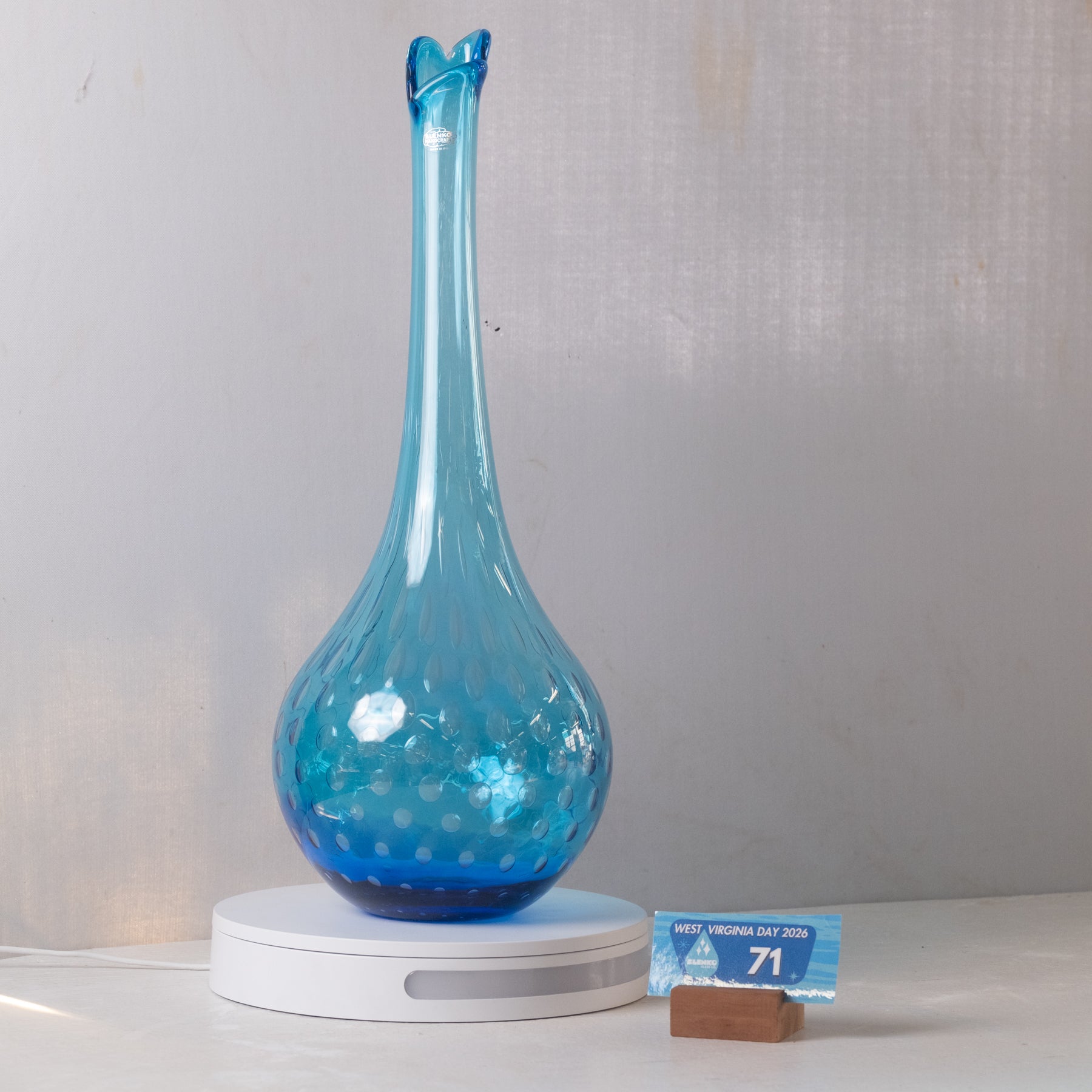 A translucent blue glass vase with a flared neck and bubble-patterned body, displayed on a white circular charging base against a light gray fabric backdrop. A small price card stands beside the base.