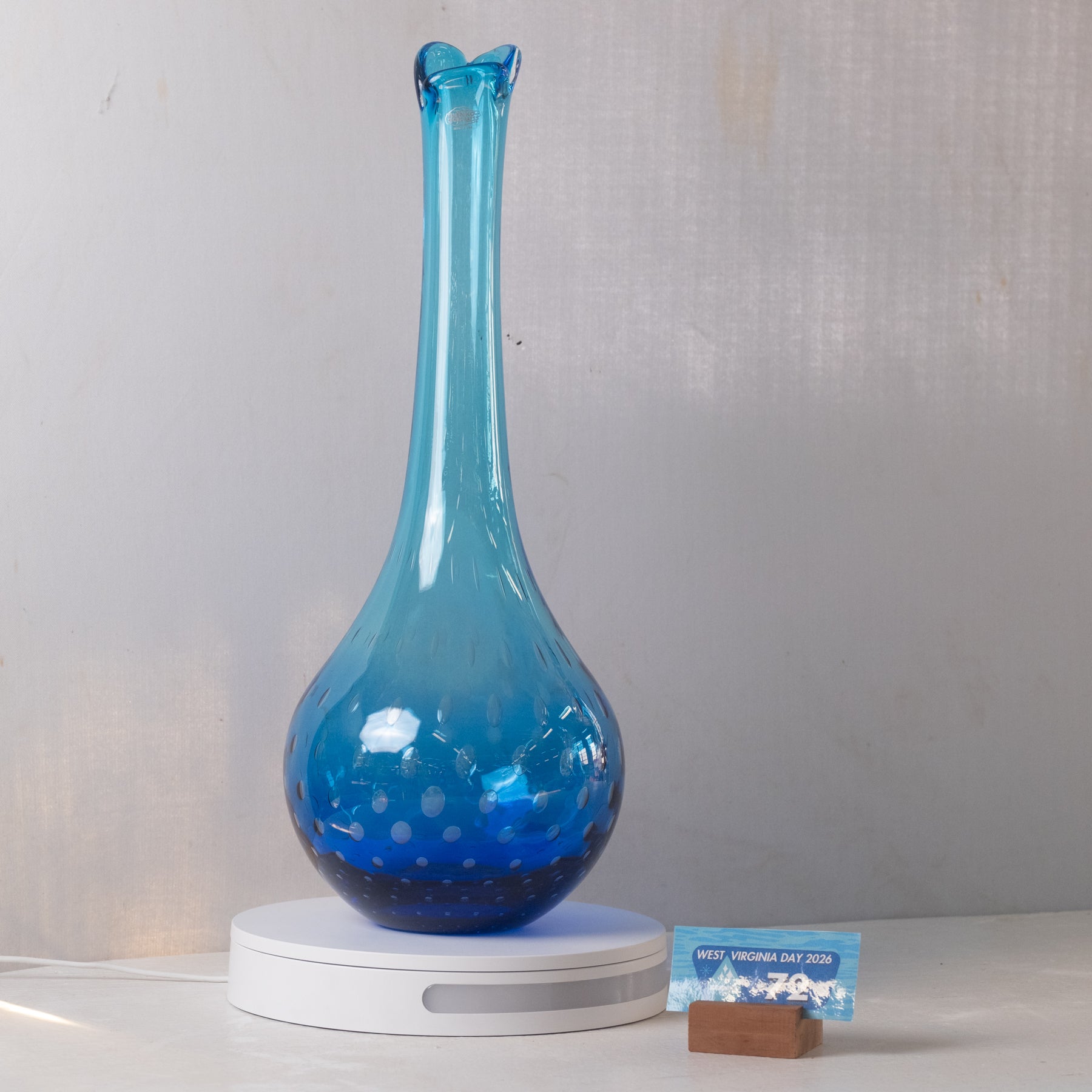 A translucent blue glass vase with a flared neck and dotted pattern, displayed on a white circular base against a light gray wall.