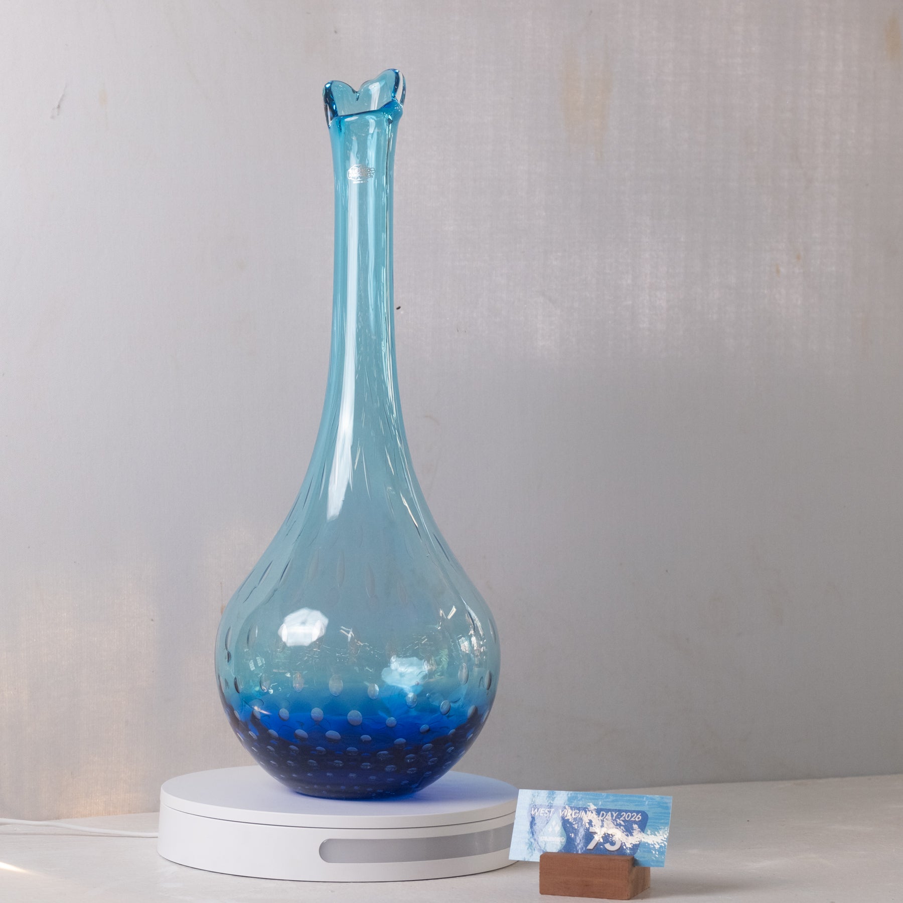 A tall, slender blue glass vase with a flared rim and textured bubble pattern, displayed on a white circular stand against a light gray wall.