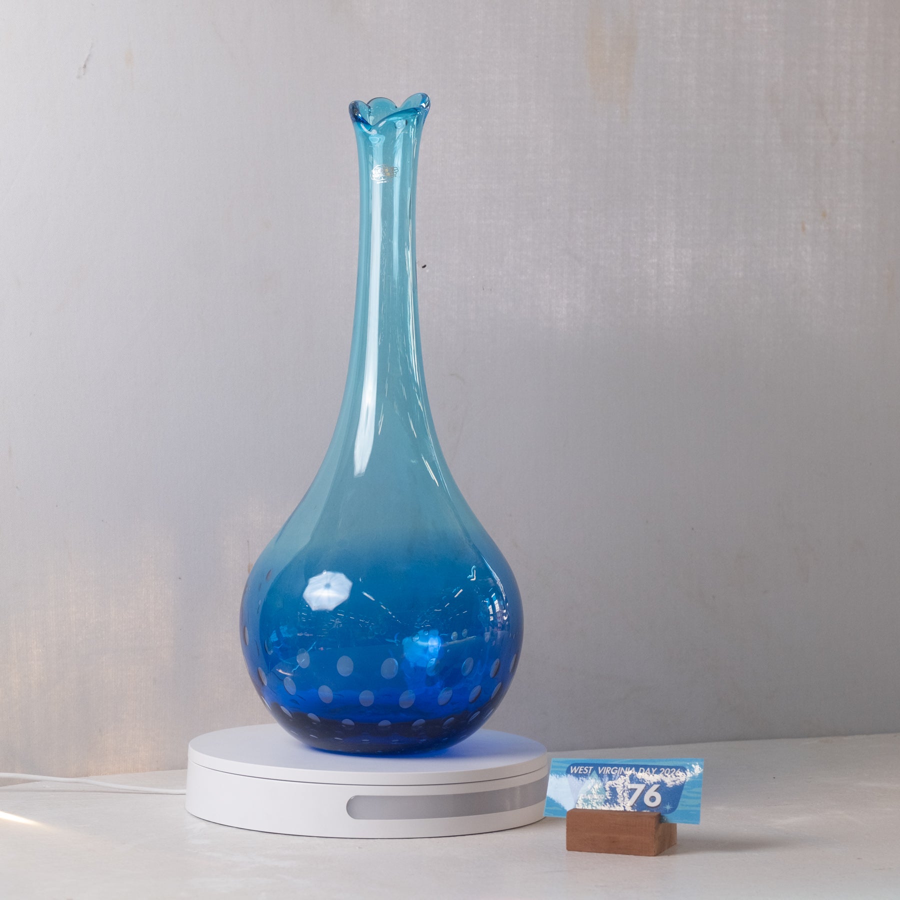 A translucent blue glass vase with a flared neck and dotted pattern near the base, displayed on a white circular stand against a light gray wall.