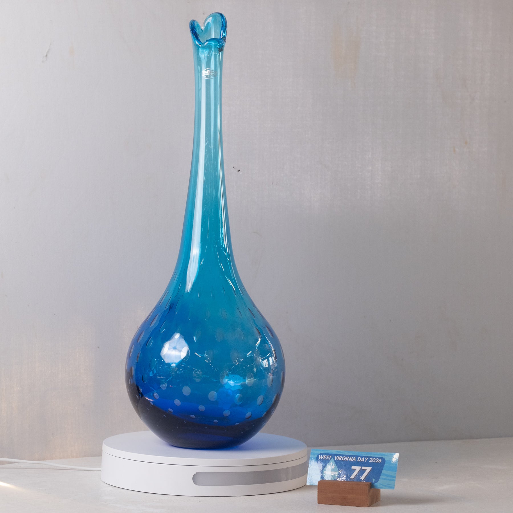 A tall, slender blue glass vase with a flared rim and dotted pattern, displayed on a white circular light box against a light gray wall.