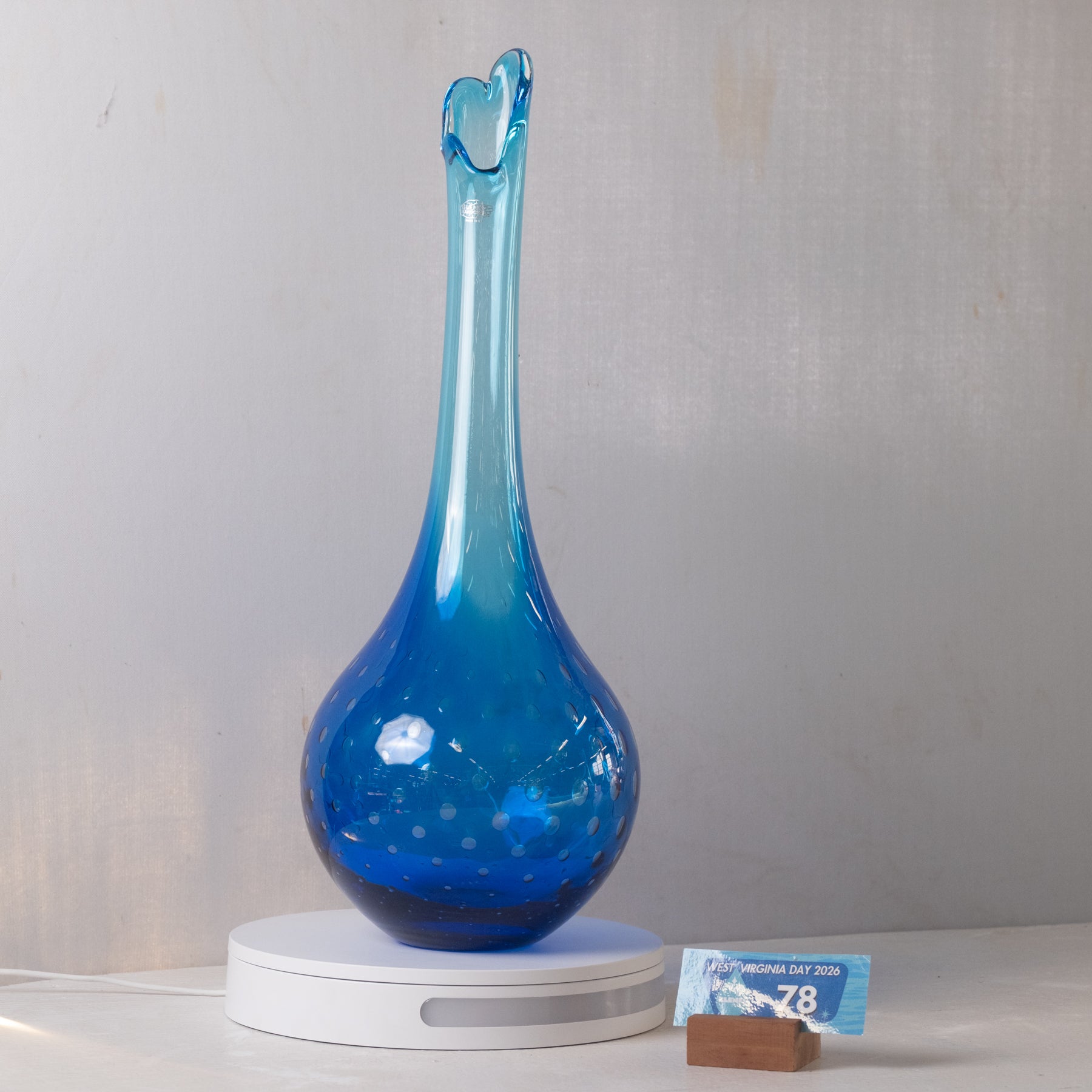 A translucent cobalt blue glass vase with a heart-shaped flared neck, displayed on a white circular lamp base next to a small packet of chocolate.