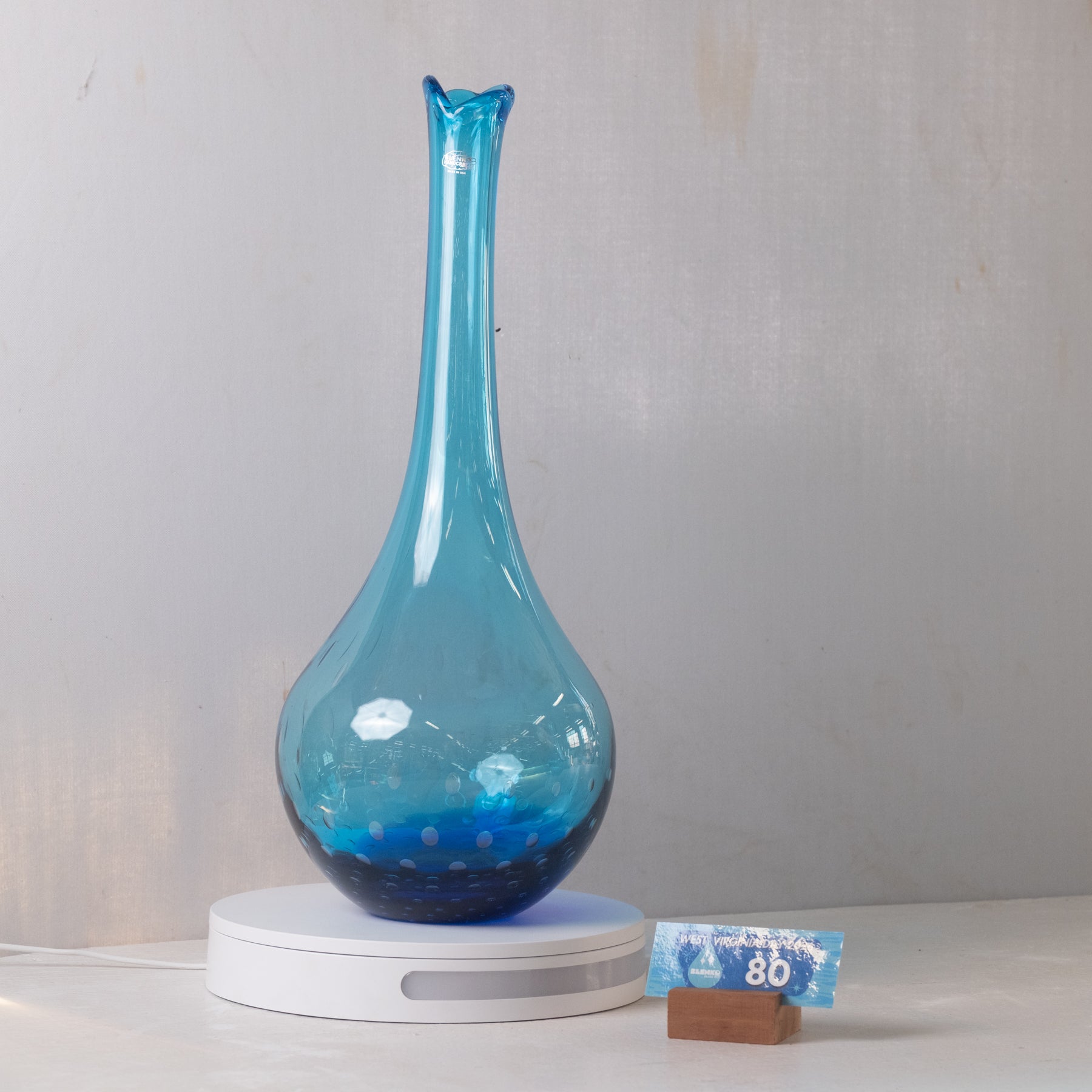 A blue glass diffuser vase with a long, flared neck and a rounded base, sitting on a white circular charging base. A small blue and white card is propped next to the base on a light-colored surface against a neutral gray wall.