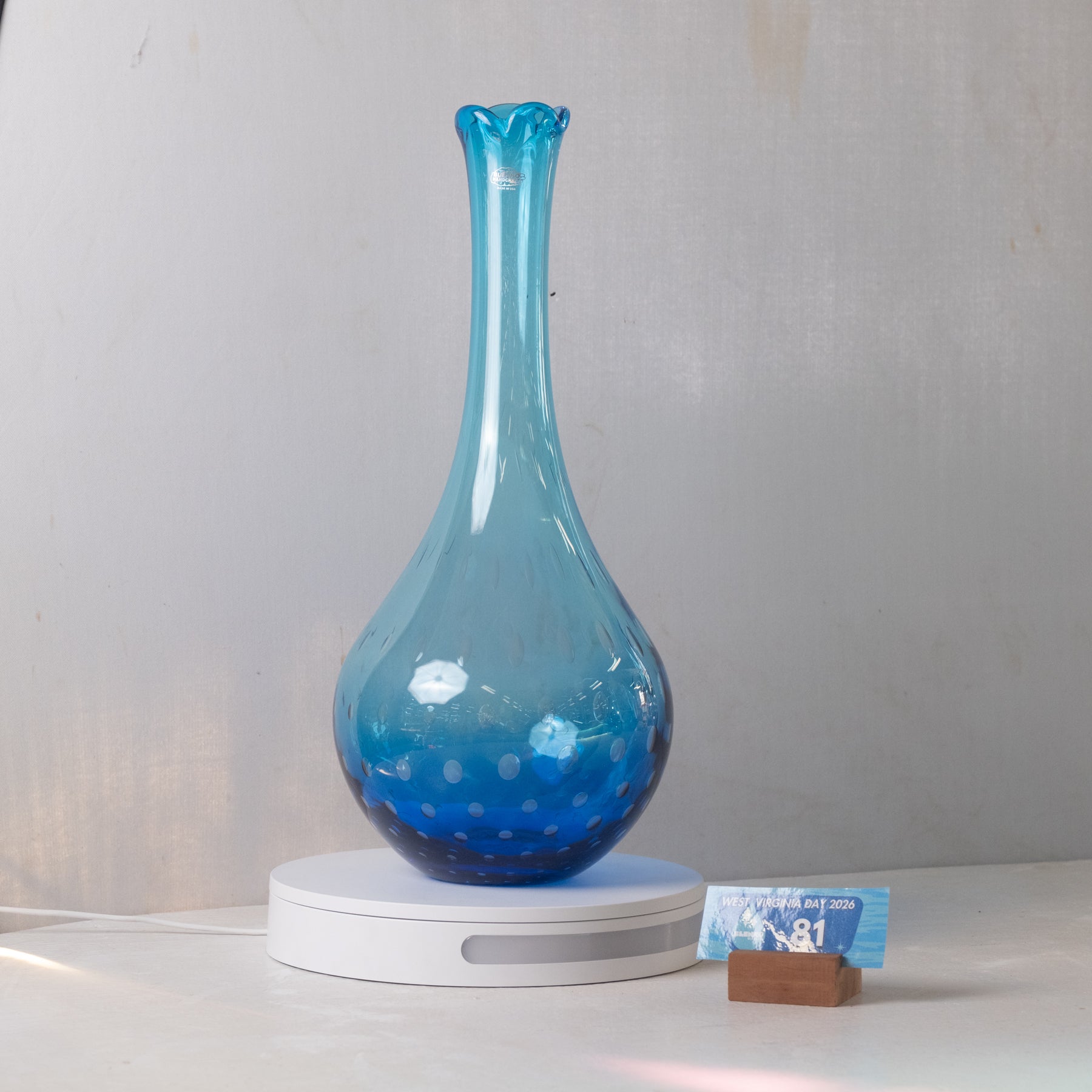 A translucent blue glass vase with a flared neck and spotted bubble pattern, displayed on a white circular LED light base with a small card in the foreground against a light gray wall.