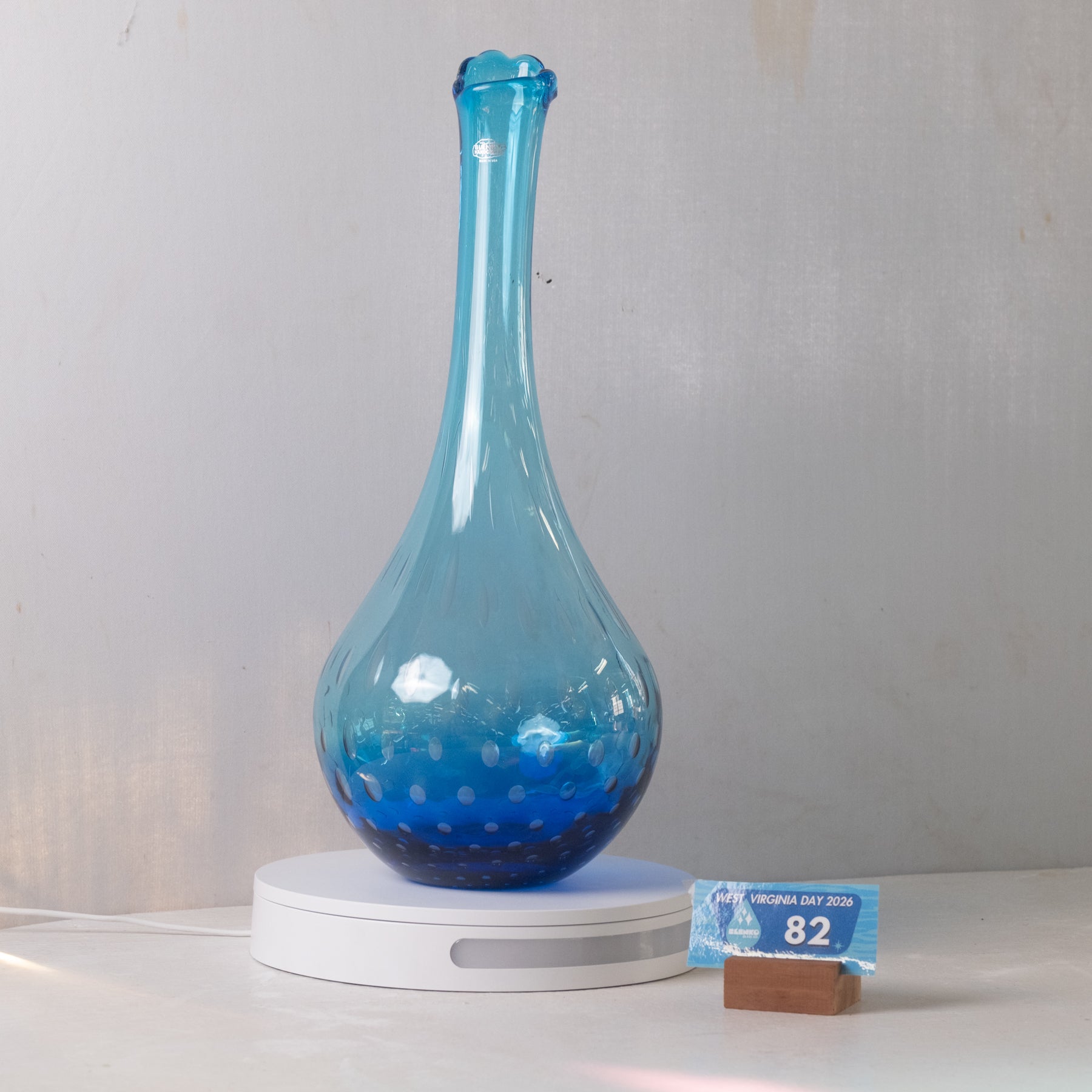 A translucent blue glass vase with a bulbous base and narrow neck, displayed on a white circular lighted base against a light gray wall. A small blue price tag card is visible at the lower right.