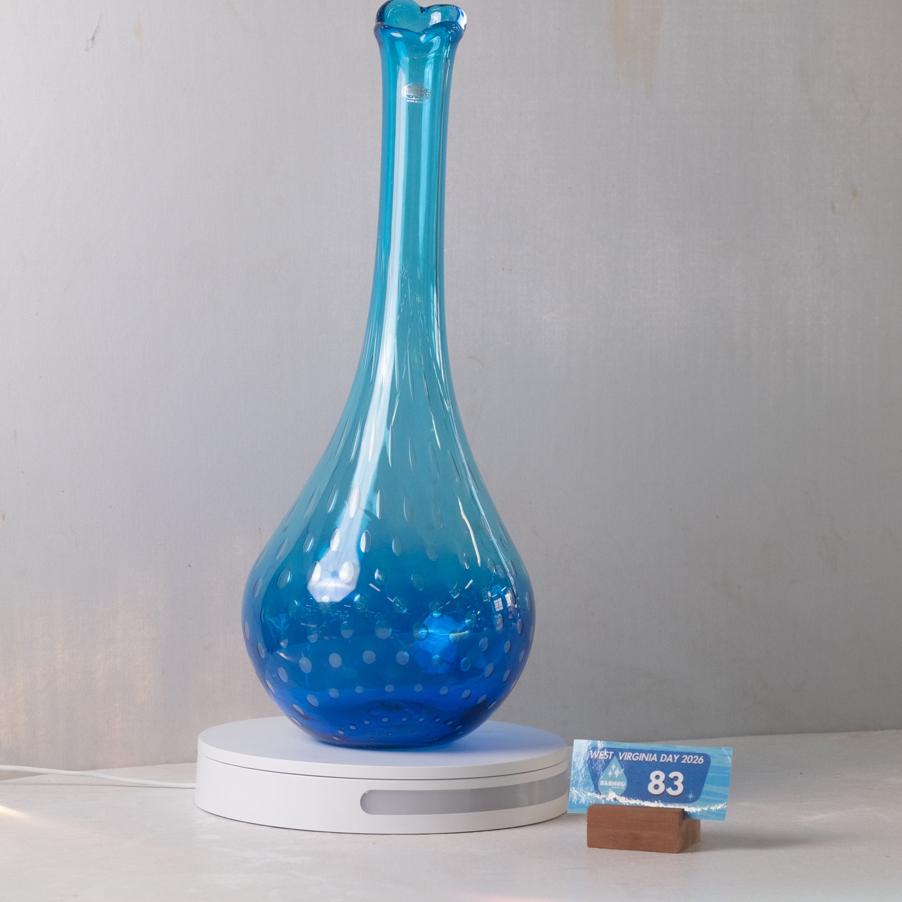 A translucent blue glass vase with a flared neck and dotted pattern, displayed on a white circular light box. A small blue and brown price tag card is visible on the right against a plain light gray wall background.