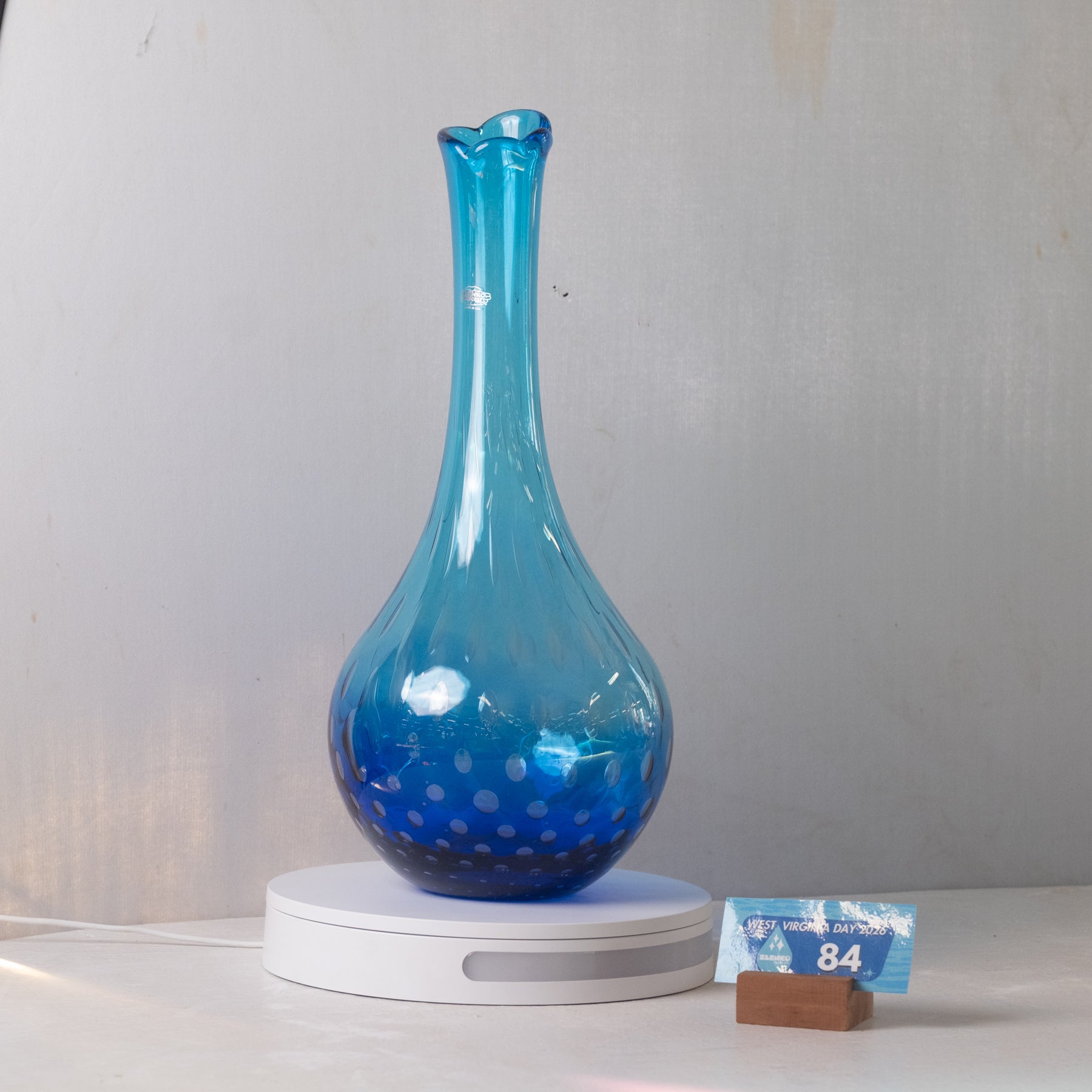 A translucent blue glass vase with a flared neck and dotted pattern near the base, displayed on a white circular light box. A small blue price tag card is resting on the right side of the platform against a plain light gray wall.