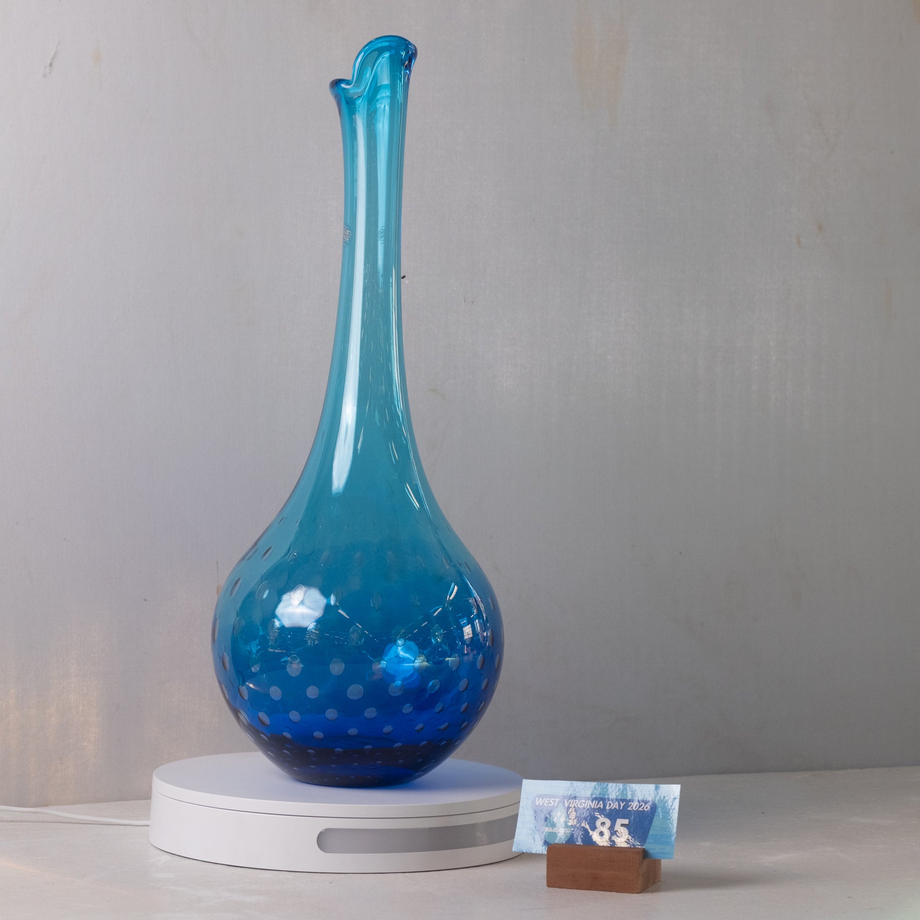 A translucent blue glass vase with a bulbous base and narrow flared neck, displayed on a white circular light box against a light gray wall. A small price card on a wooden stand sits beside the base.