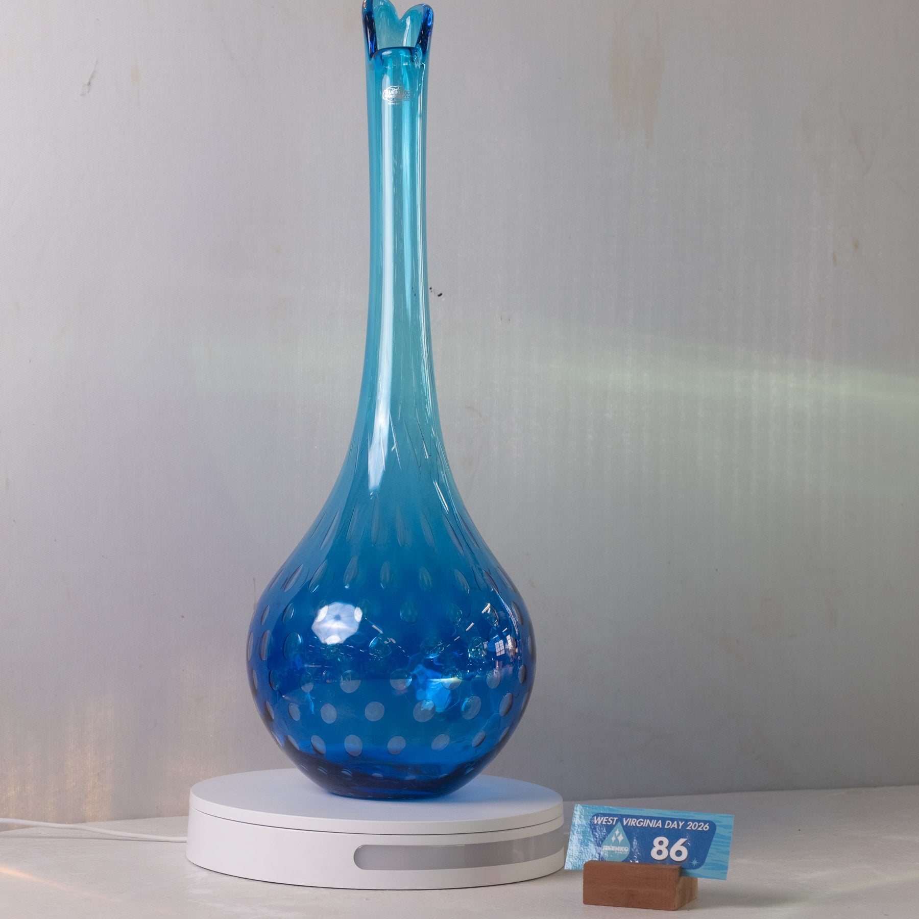 A blue glass vase with a long neck and dotted pattern, displayed on a white circular base next to a small price card.