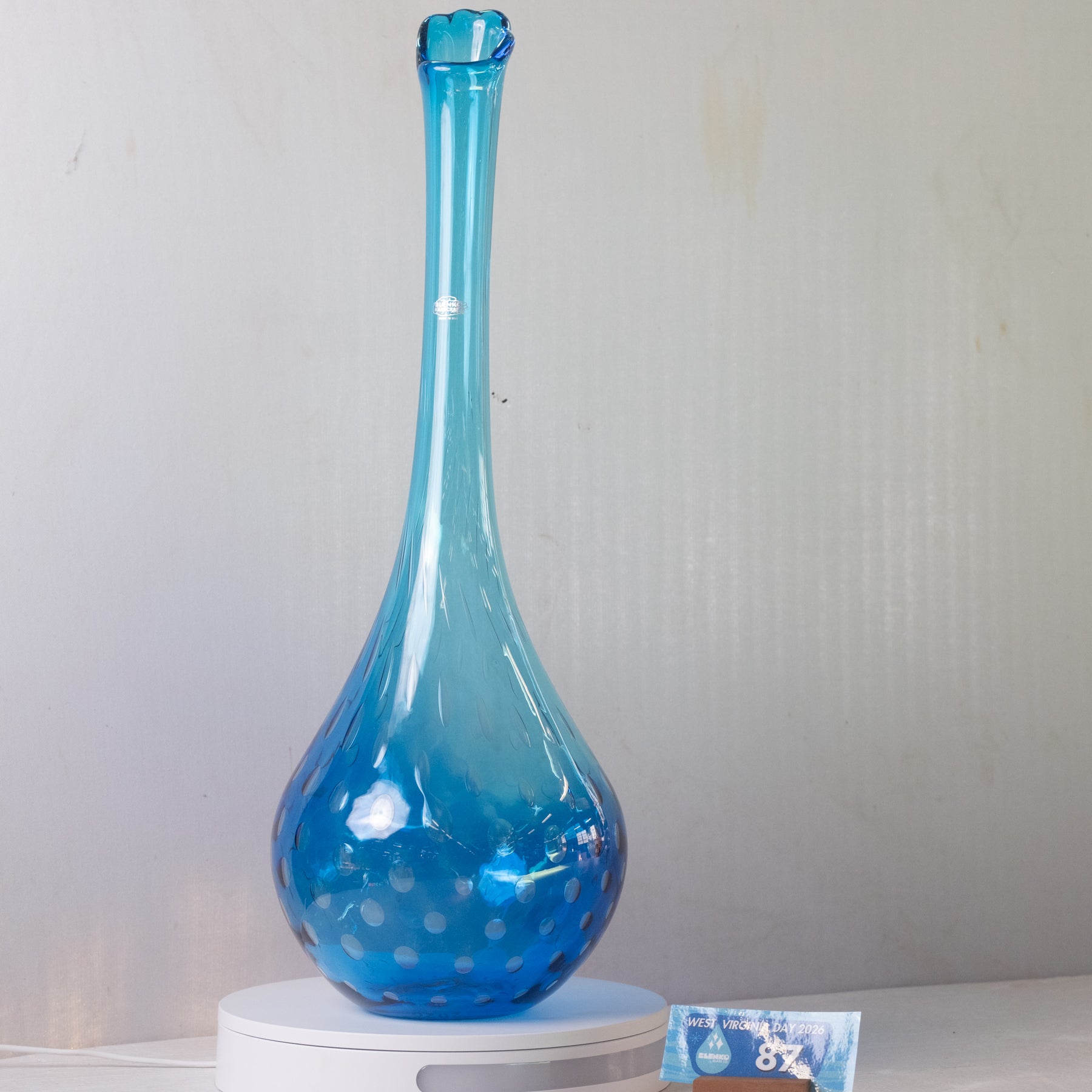 A translucent blue glass vase with a bulbous base and narrow neck, displayed on a white circular scale against a light gray wall.