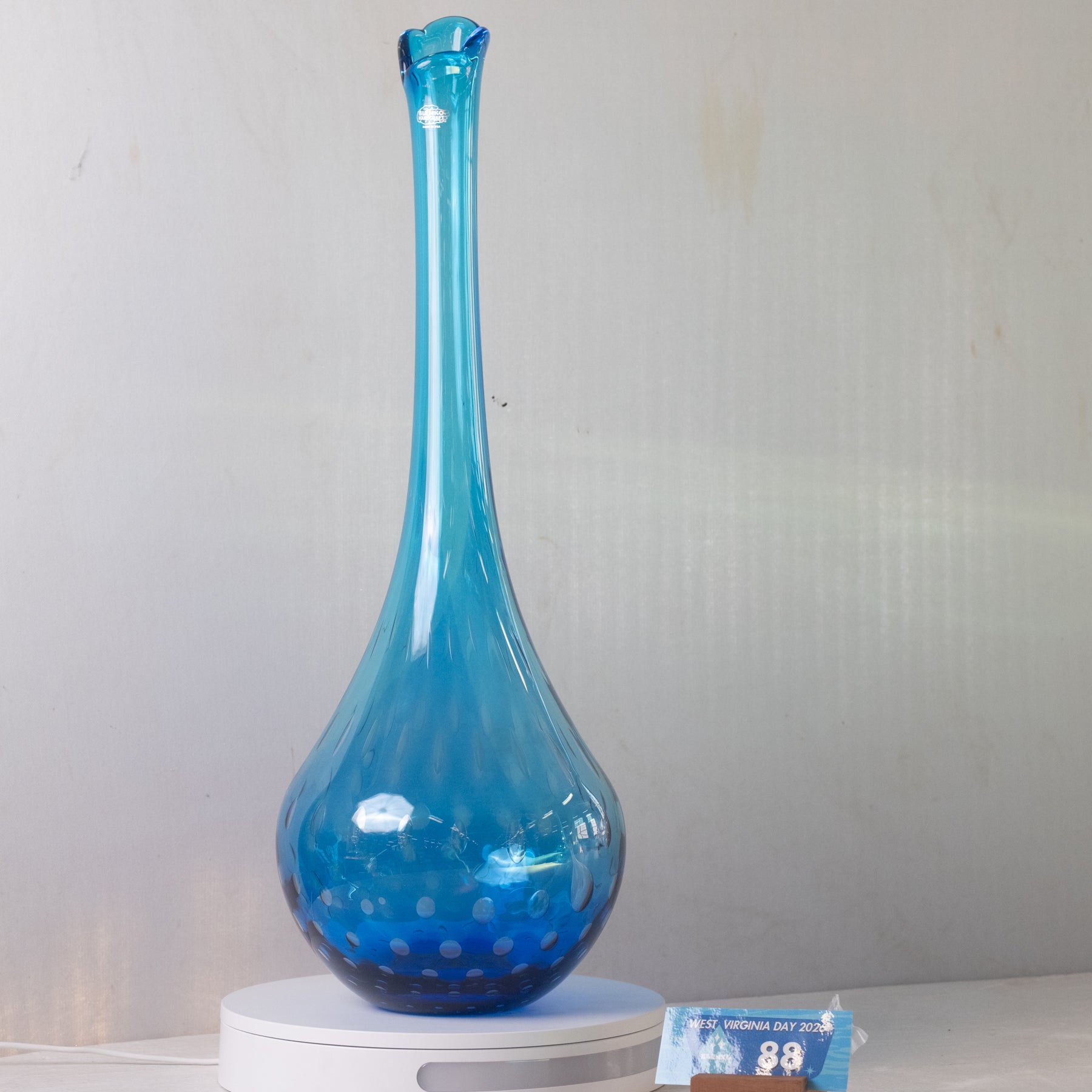 A tall, slender blue glass vase with a bulbous base and small bubble-like speckles, displayed on a white round stand against a light gray wall.