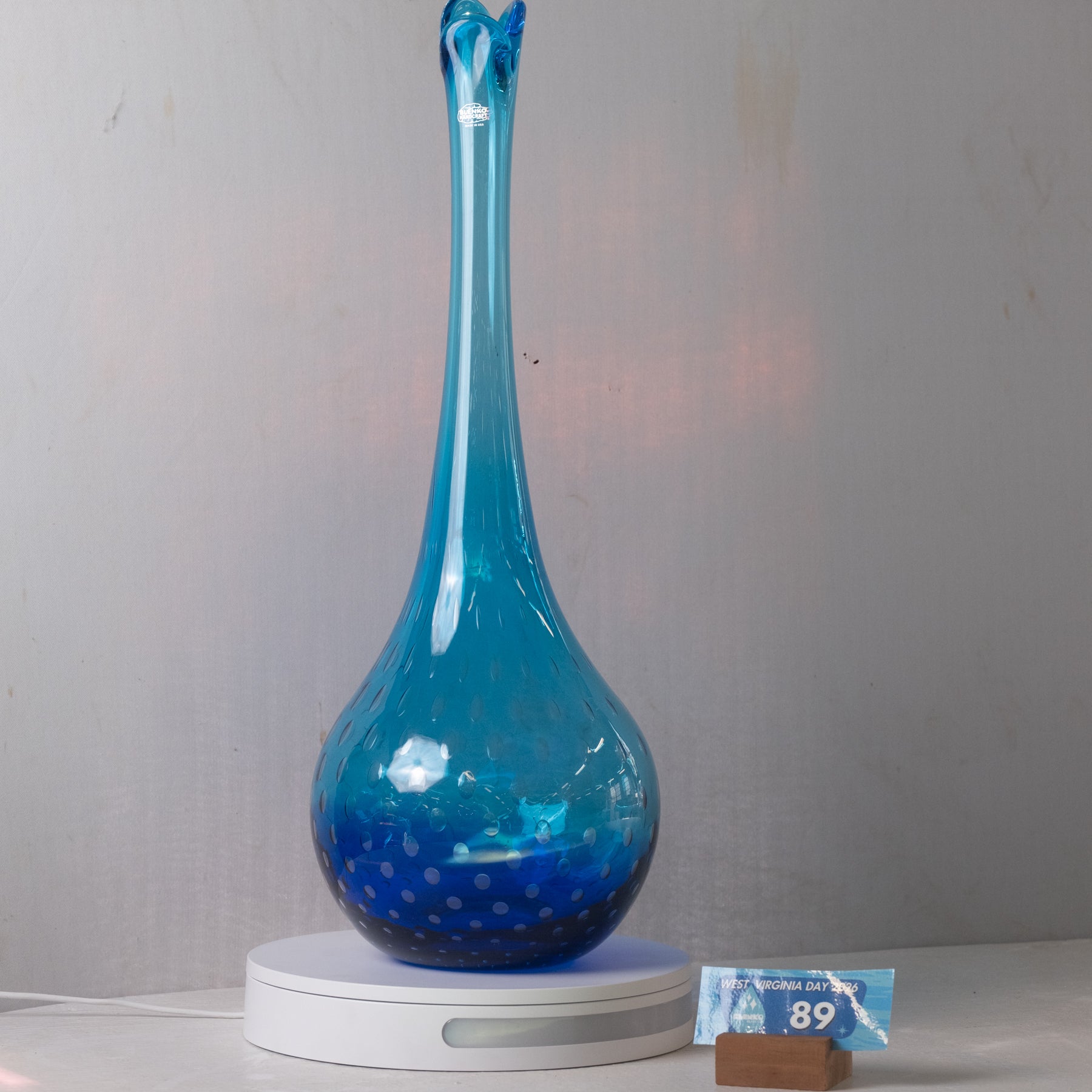 A tall, narrow blue glass vase with a bulbous base and small white speckles, displayed on a white circular light box against a plain gray wall.