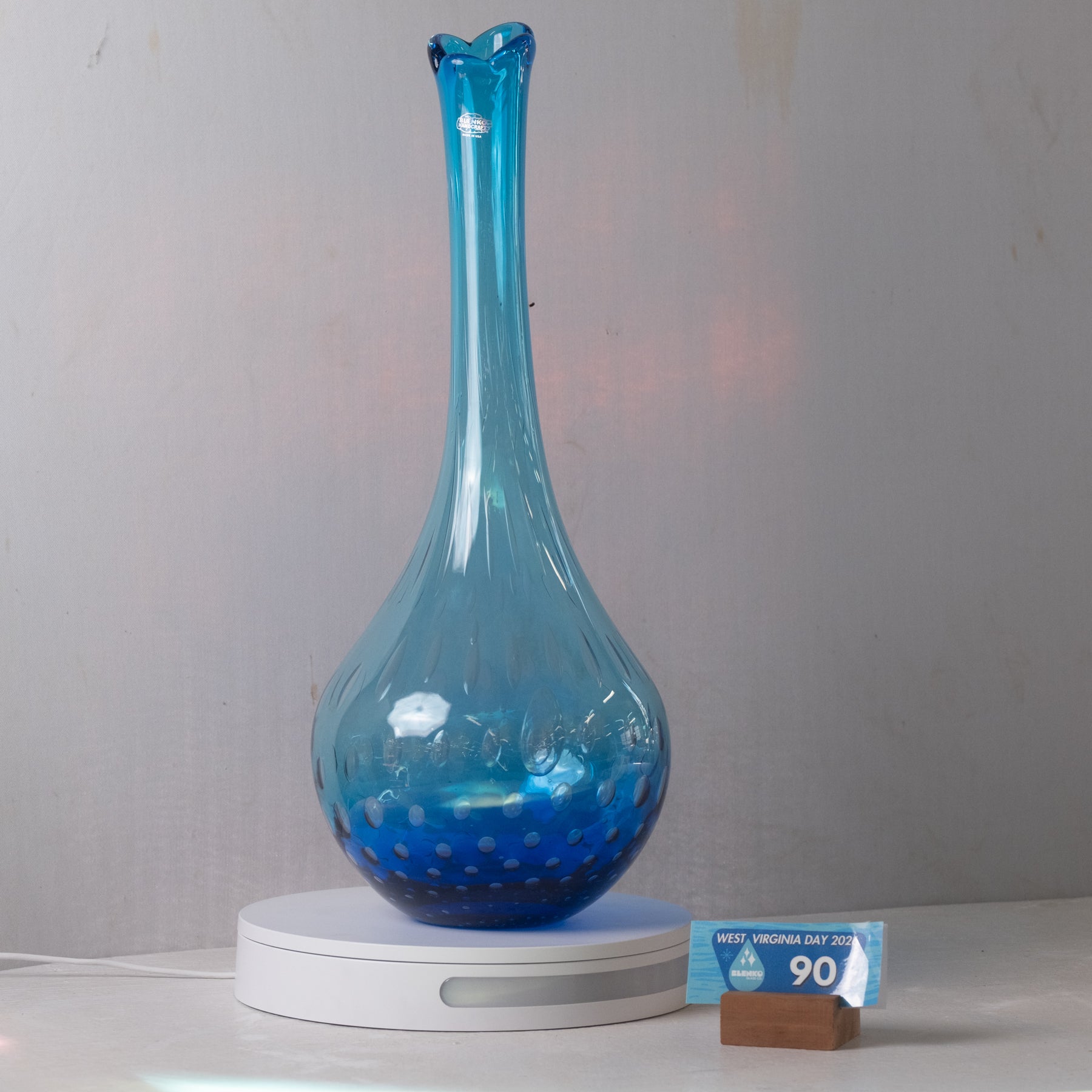 A blue glass vase with a flared neck and dotted pattern near the base, displayed on a white circular light box. A small blue and brown price tag card is visible on the right side against a plain gray wall background.