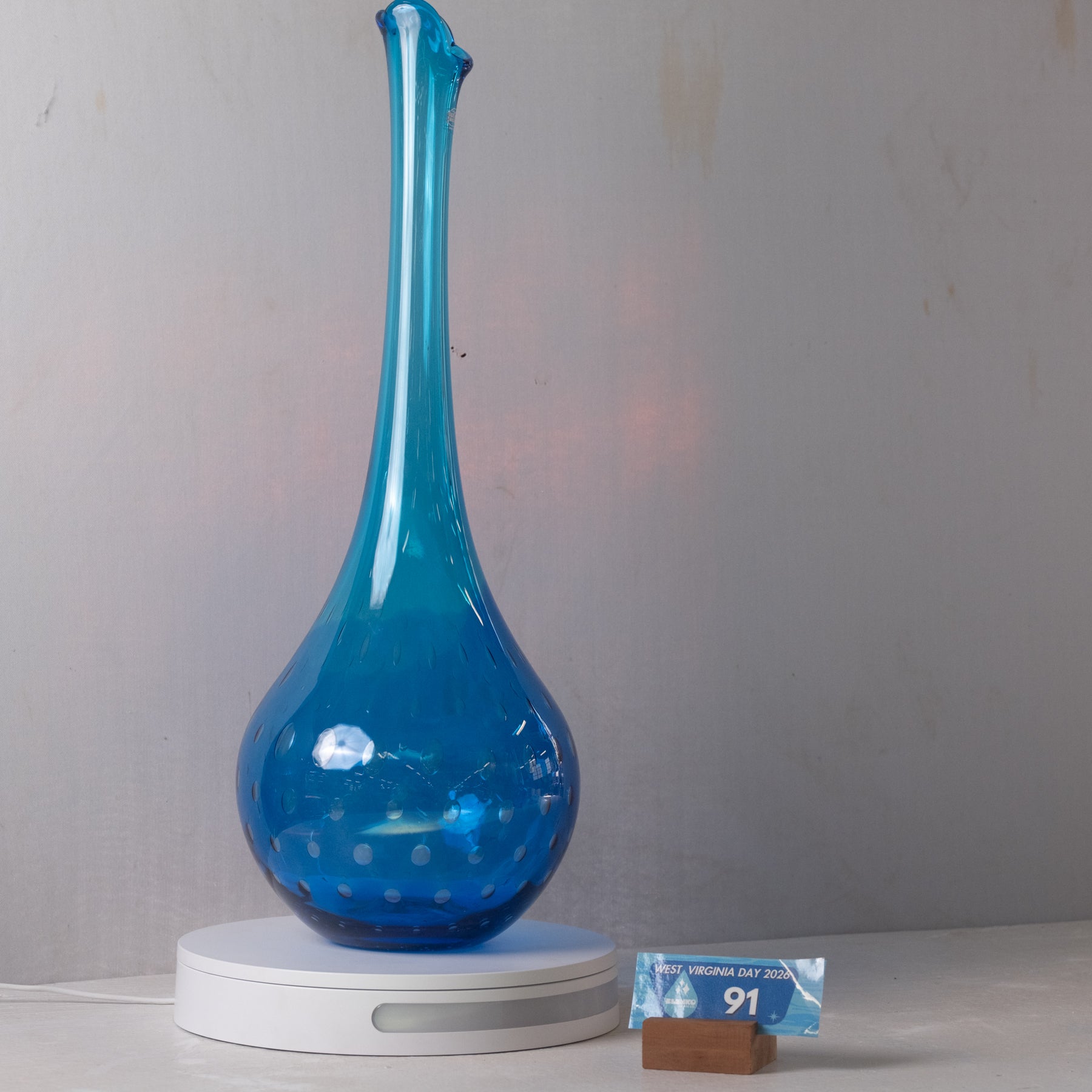 A translucent cobalt blue glass vase with a flared neck and small white polka dots near the base, displayed on a white circular lamp base against a light gray wall.