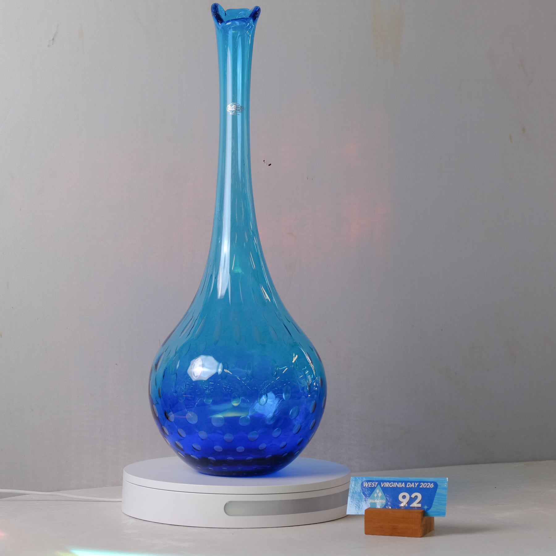 A blue glass perfume bottle with a flared neck and perforated base, displayed on a white circular stand against a light gray wall. A small blue card and wooden block sit beside the stand.