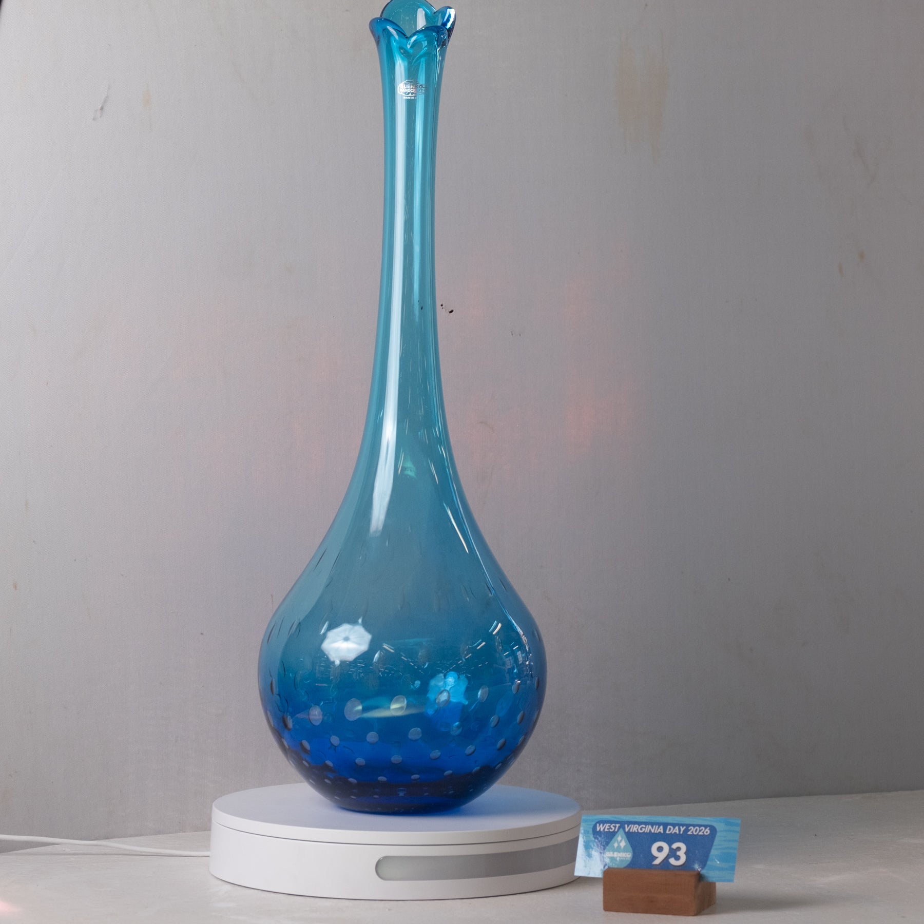 A tall, bulb-shaped blue glass vase with a flared neck sits on a white circular base. The vase has a glossy finish with small white speckles near the bottom.