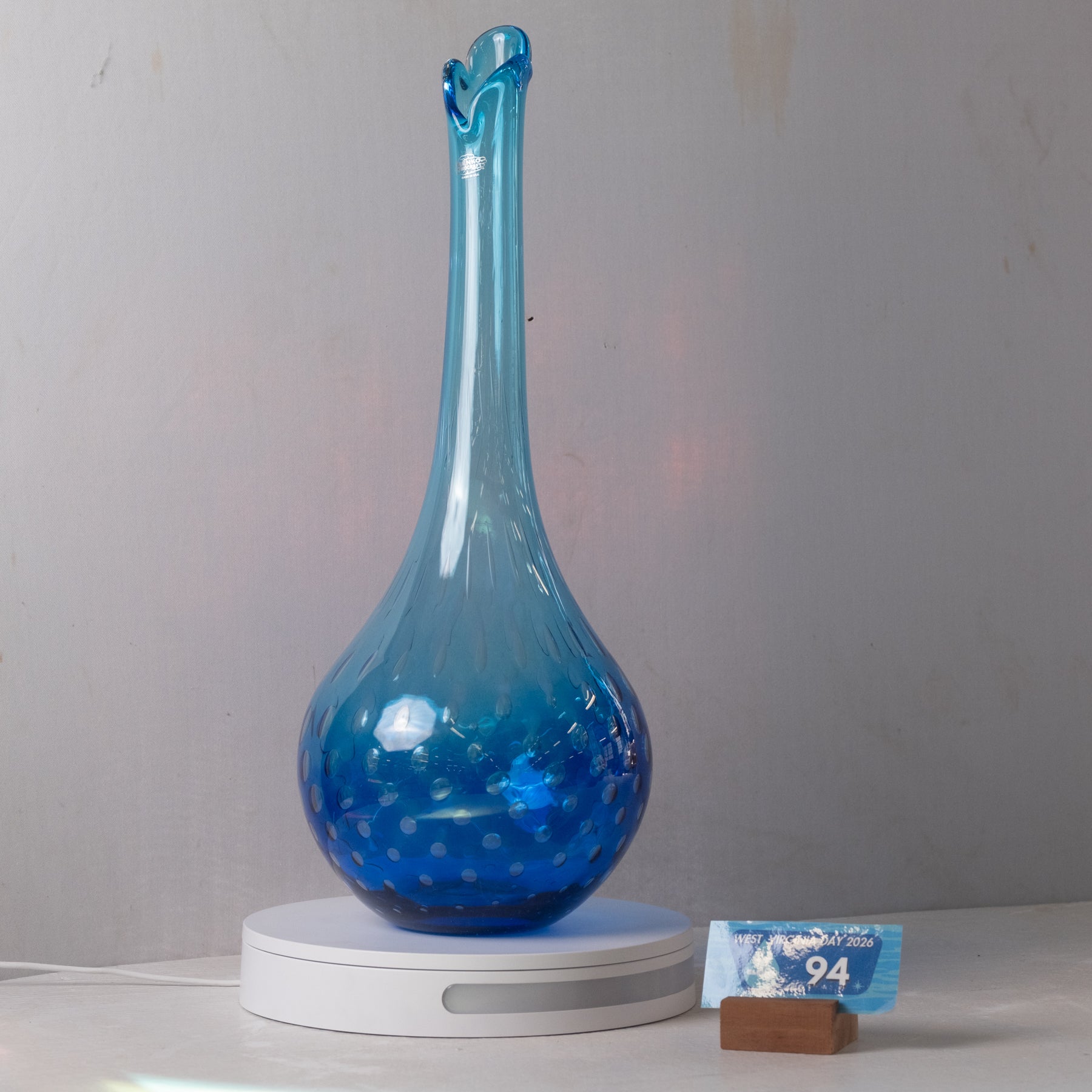 A translucent blue glass vase with a flared neck and dotted pattern, displayed on a white circular base against a plain light gray wall.