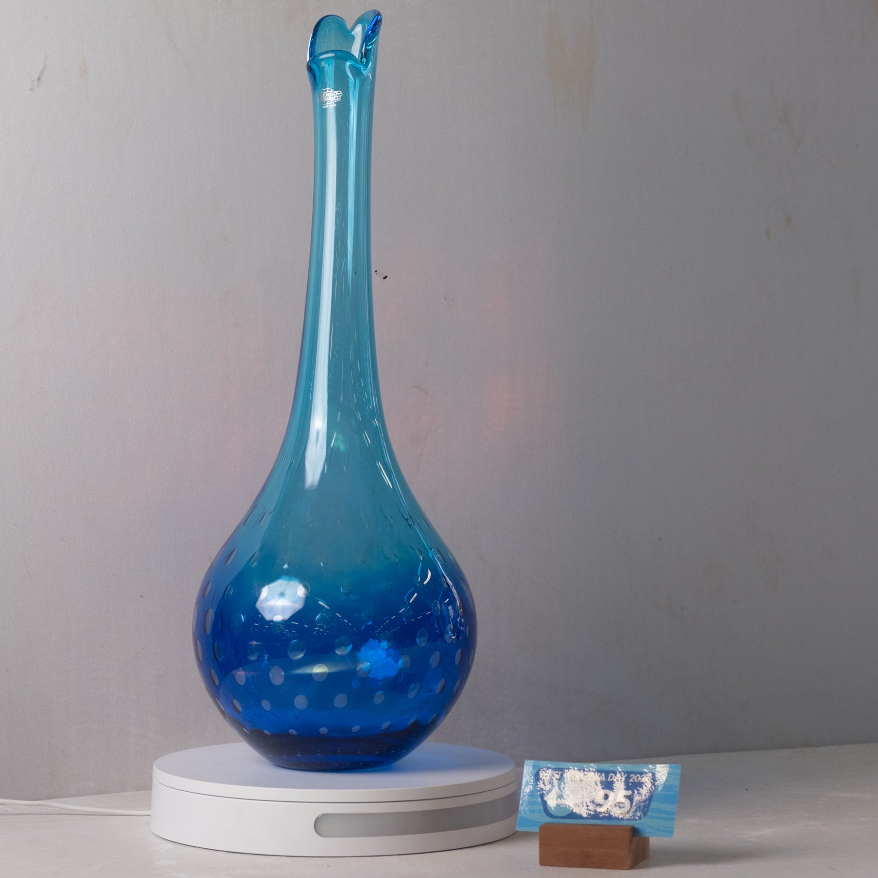 A translucent blue glass vase with a flared neck and small loop at the top, resting on a white circular base. The vase has a gradient from lighter sky blue at the top to deep cobalt blue at the bottom, with small white polka dots near the base.