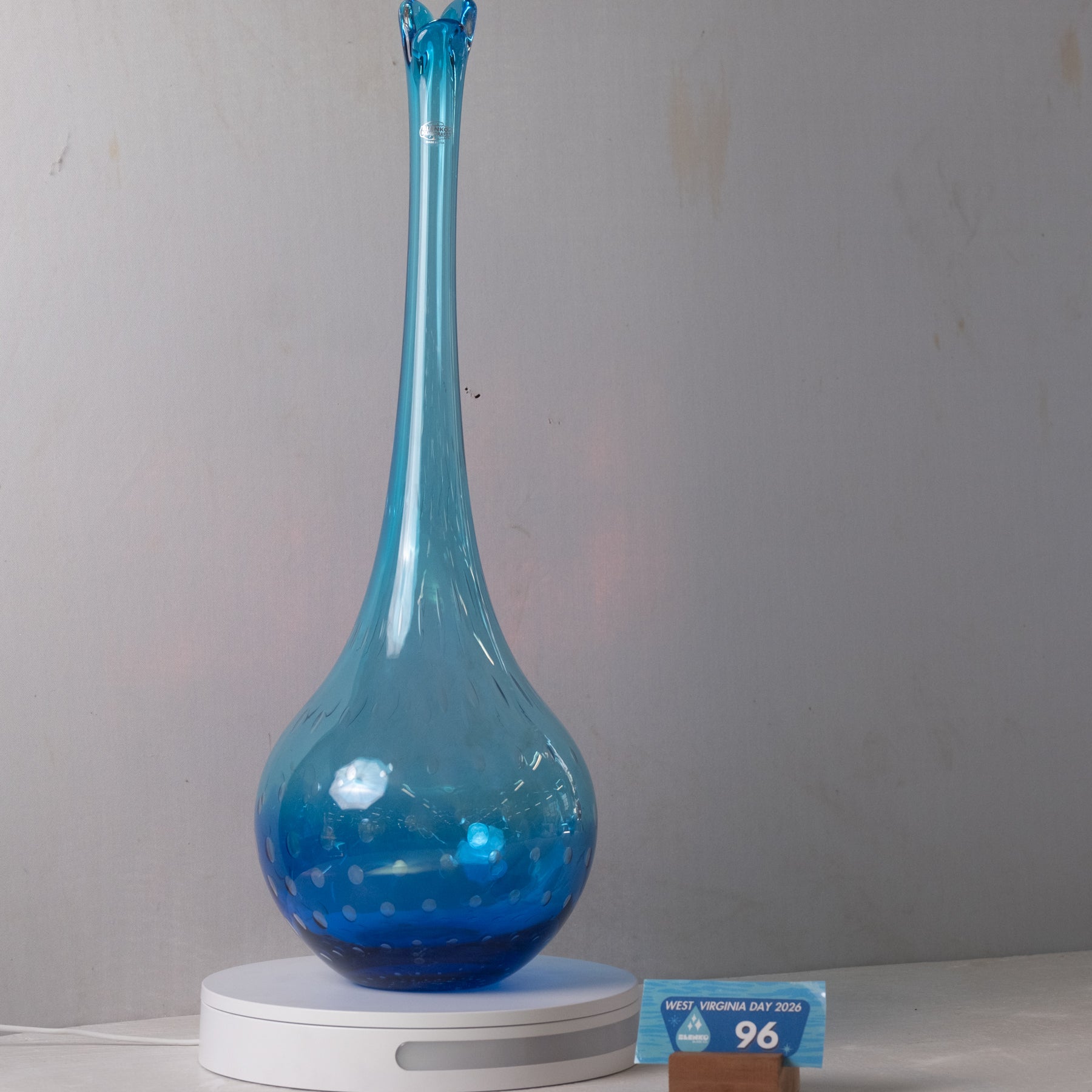 A tall, slender blue glass vase or lamp base with a bulbous rounded body, displayed on a white circular base against a plain light gray wall. A small blue price tag is visible at the lower right.