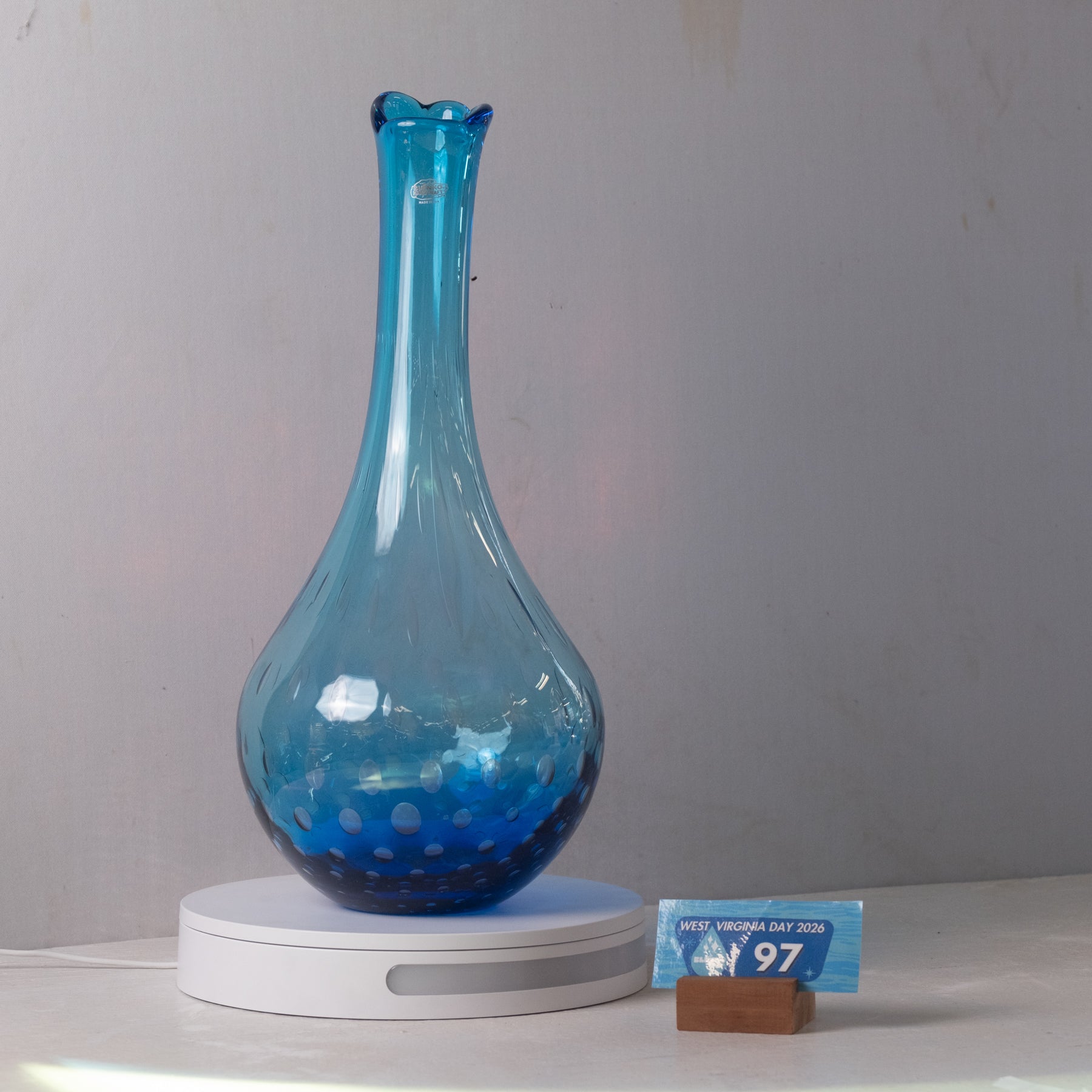 A translucent blue glass vase with a flared rim and subtle white speckled pattern near the base, displayed on a white round scale with a small price tag in front, against a plain light gray wall.