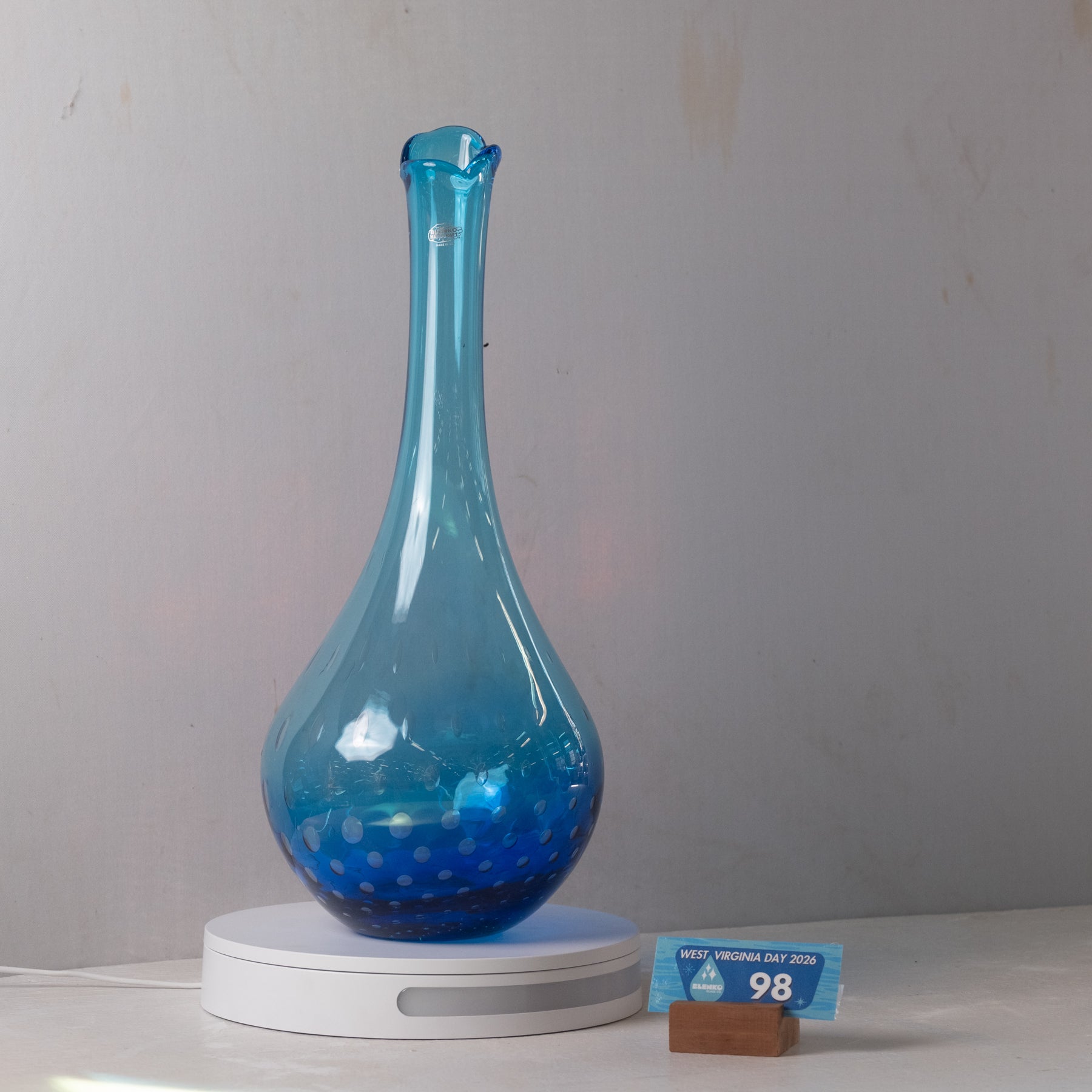 A translucent blue glass vase with a flared neck and dotted textured base, displayed on a white circular lighted base against a light gray wall. A small blue size card on a wooden block shows 98 cm.