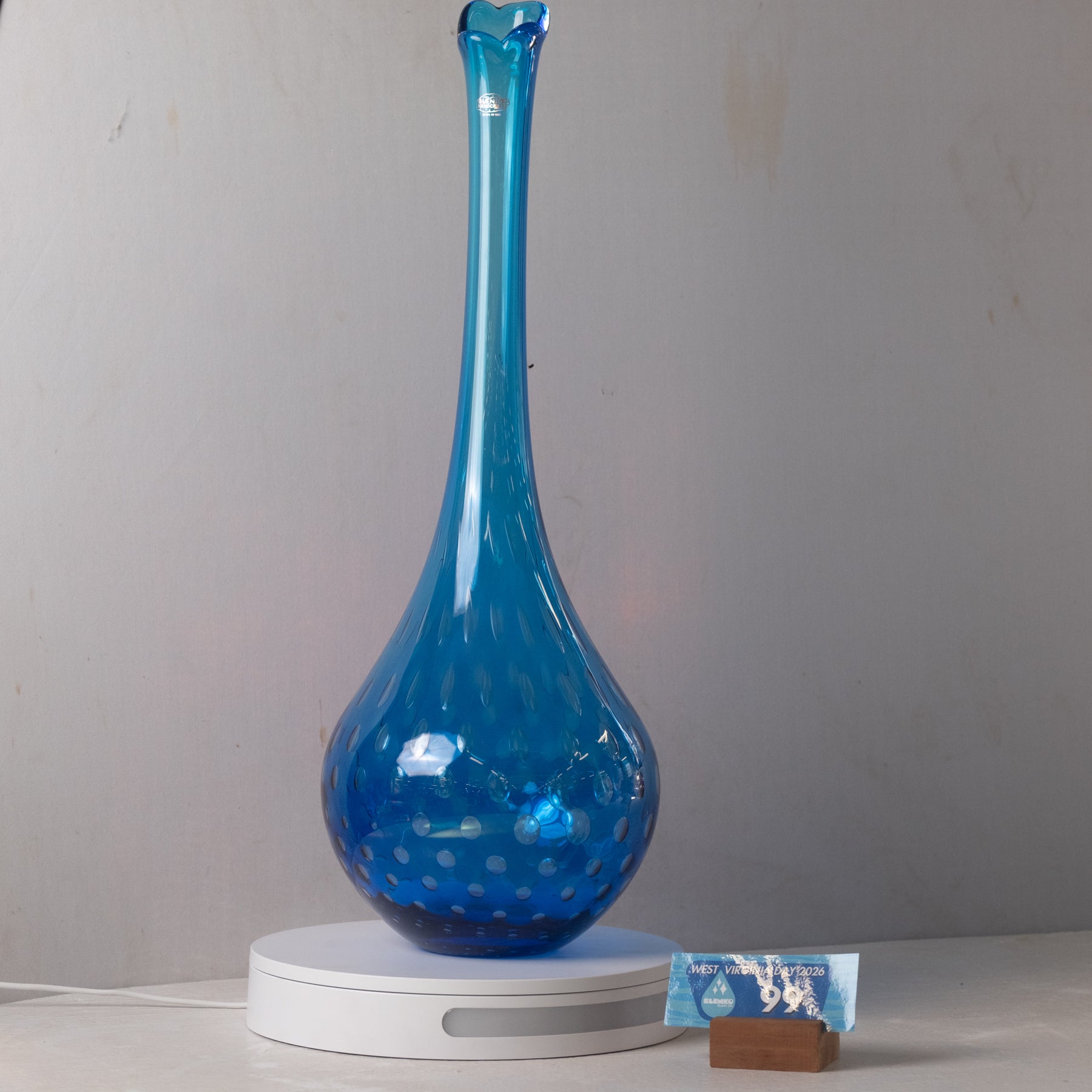 A tall, slender cobalt blue glass vase with a flared neck and small white polka dots, displayed on a white circular light box against a plain light gray wall.