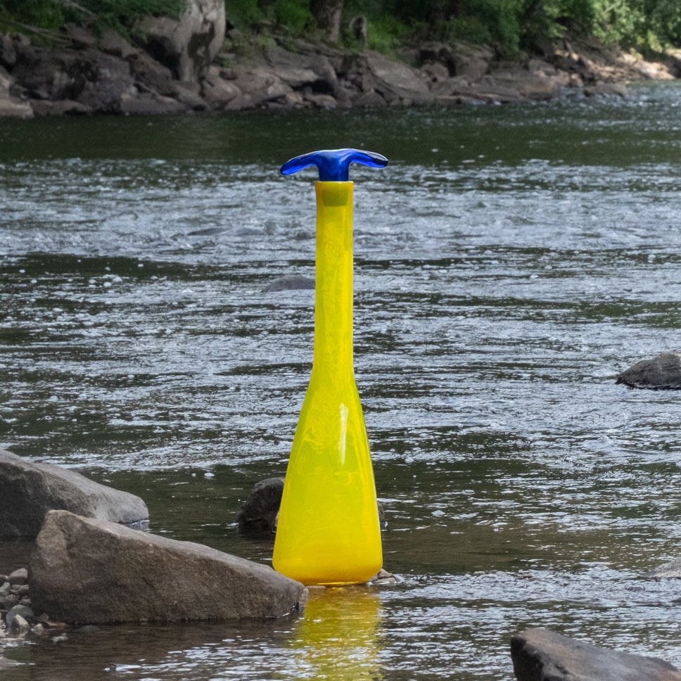 Yellow bottle with blue cap floating in a river with a bridge and trees in the background