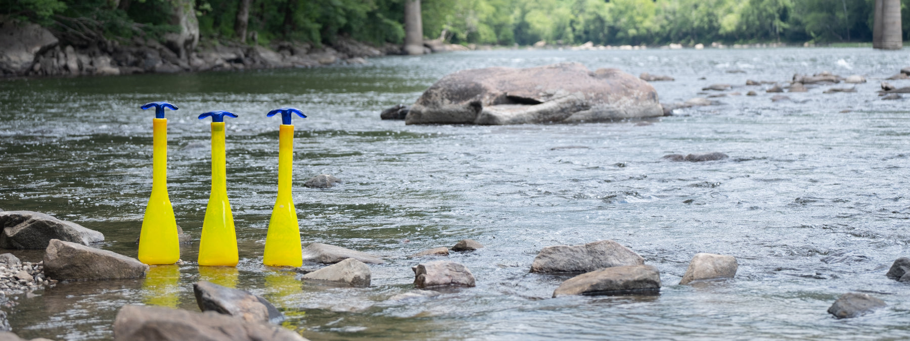 Three yellow buoys with blue tops floating in a river with rocks and trees in the background.