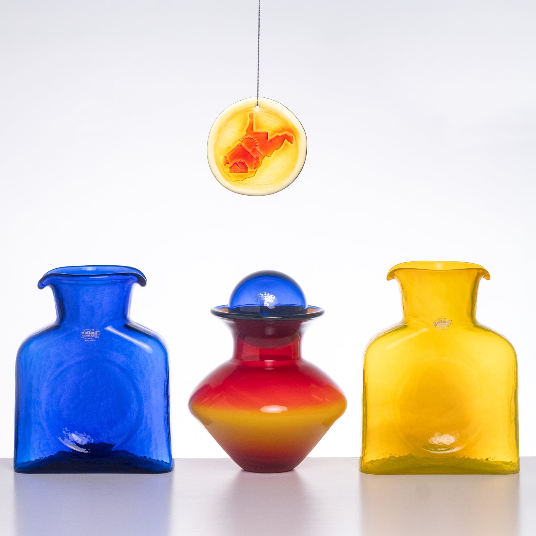 Three colorful glass vases on a white surface with a hanging yellow and orange abstract sculpture in the background.