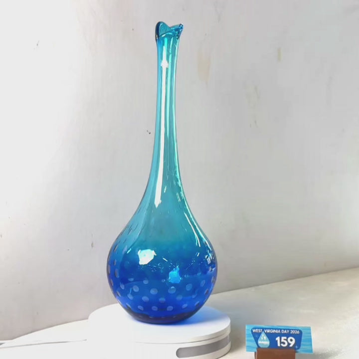 A translucent blue glass vase with a flared rim and dotted pattern near the base, displayed on a white circular stand against a plain light gray wall.