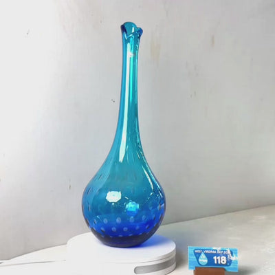 A translucent blue glass vase with a flared neck and dotted pattern near the base, displayed on a white circular light box. A small wrapped item and a brown block are placed beside it on a white surface against a plain light gray wall.
