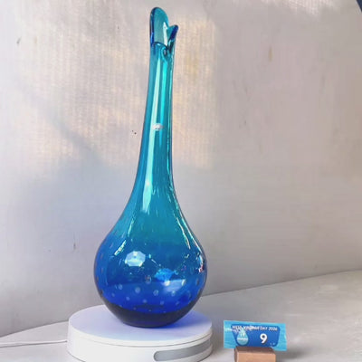 A translucent blue glass vase with a flared neck and dotted pattern, displayed on a white circular base against a light gray wall.