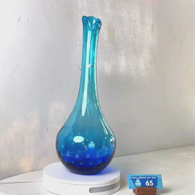 A translucent blue glass vase with a flared neck and dotted pattern, displayed on a white circular base against a light gray wall.